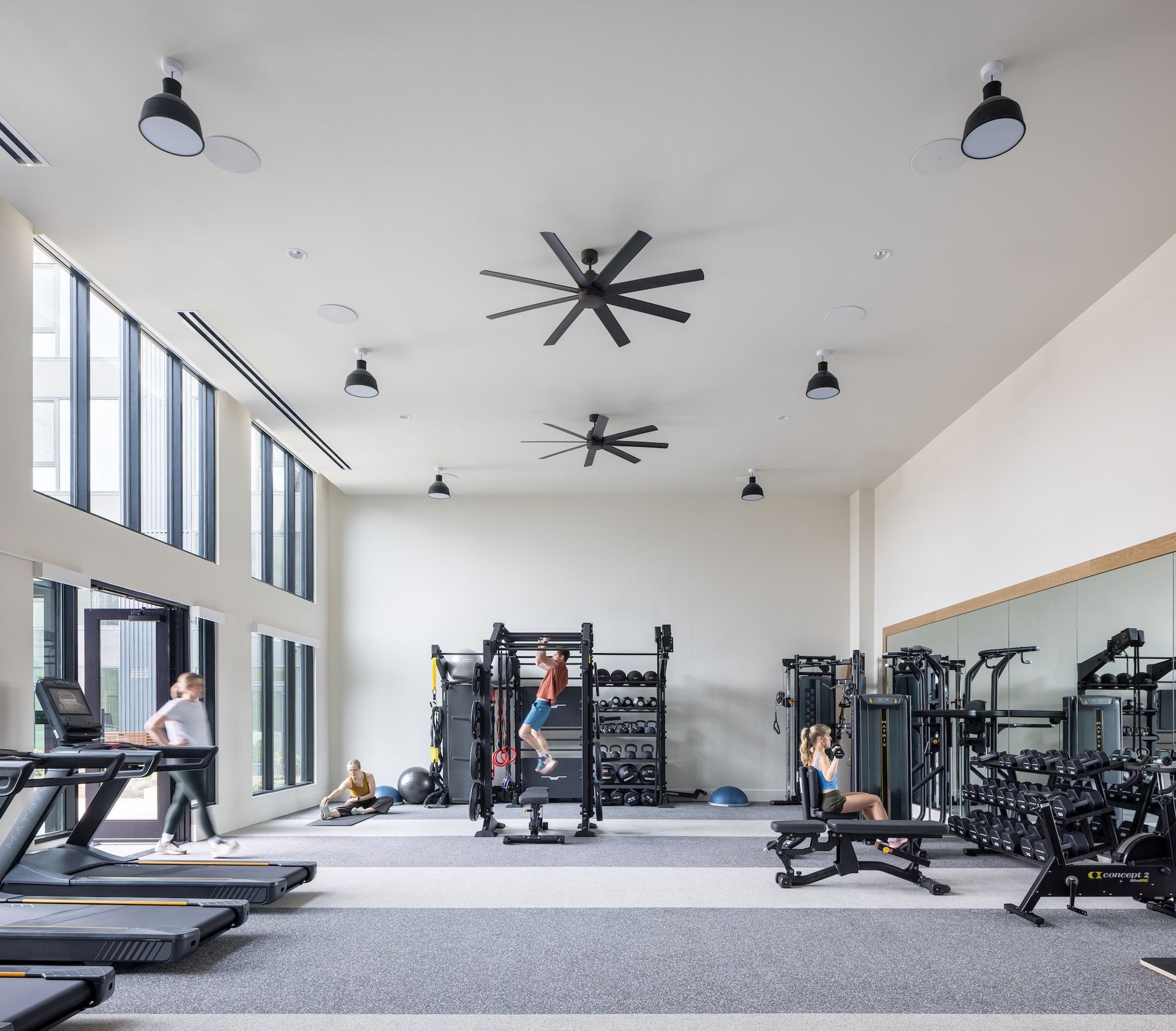 People exercising in gym with large windows. Various workout equipment, including treadmills, and weights.