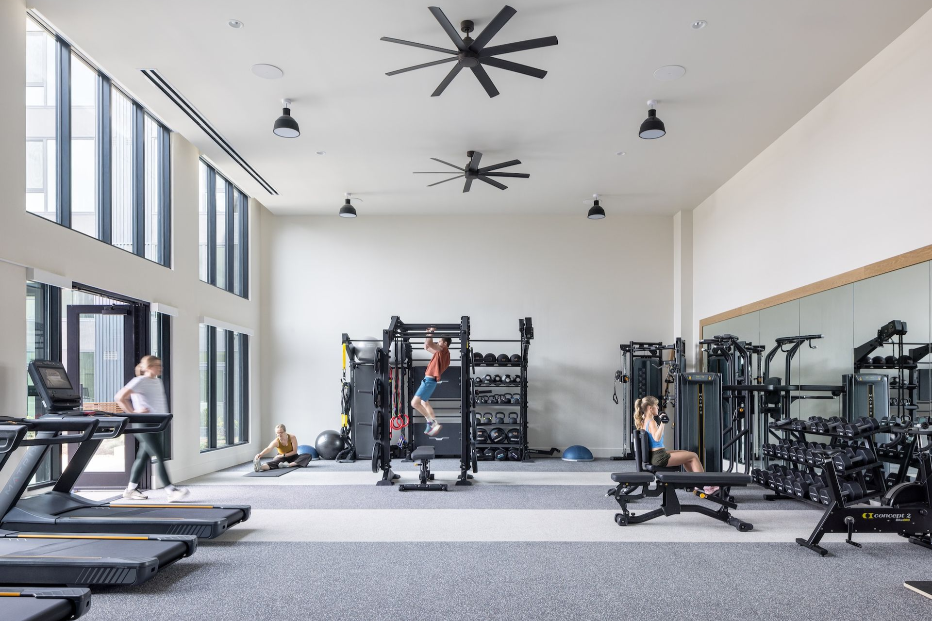People exercising in gym with large windows. Various workout equipment, including treadmills, and weights.