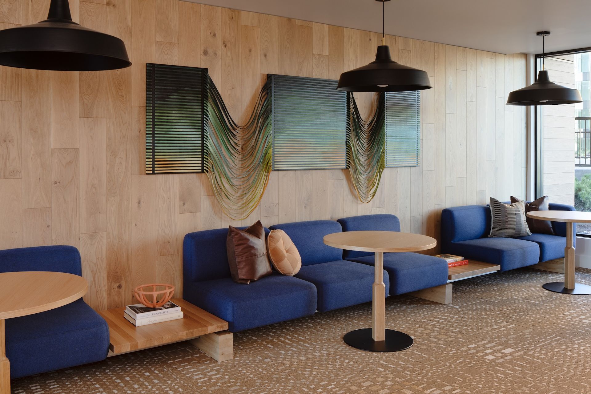Cozy lounge with blue sofas, round tables, and art on a wooden wall. Black pendant lights hang overhead.