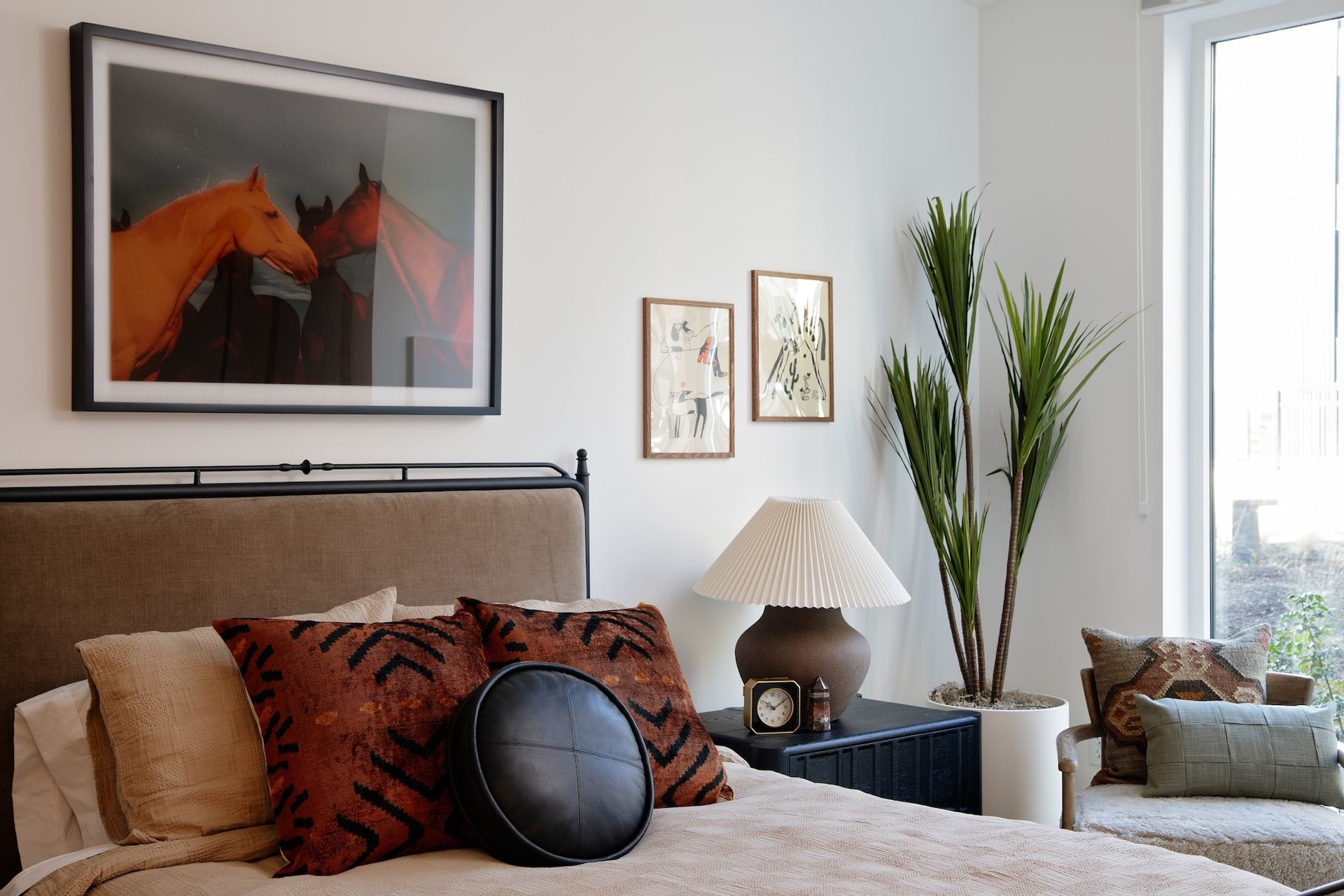 Bedroom with bed, horse art, lamp, and tall plant next to a window with seating.