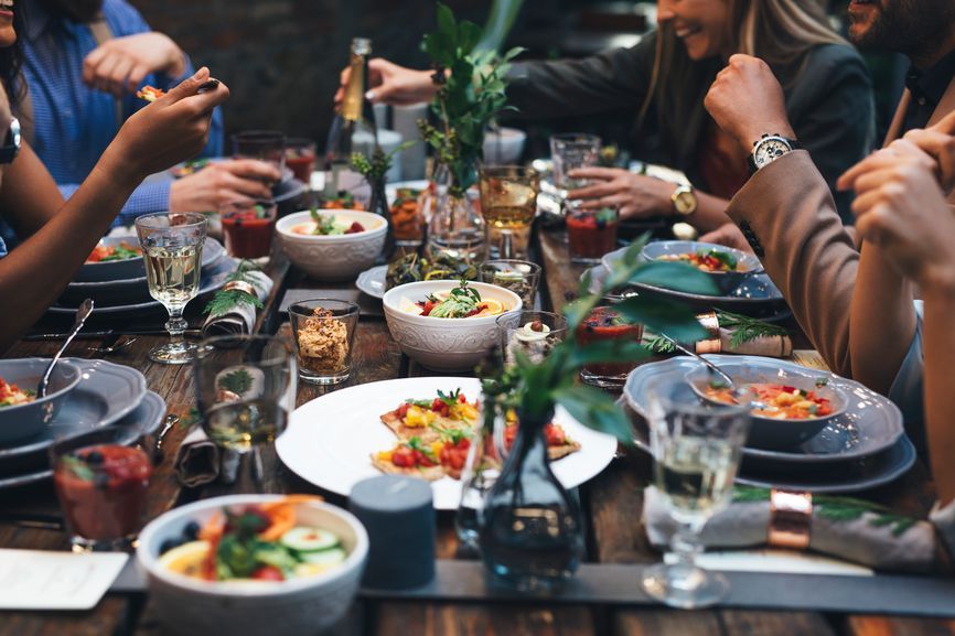 People seated at a table, enjoying a meal. Dishes of food, drinks, and floral centerpieces are present.