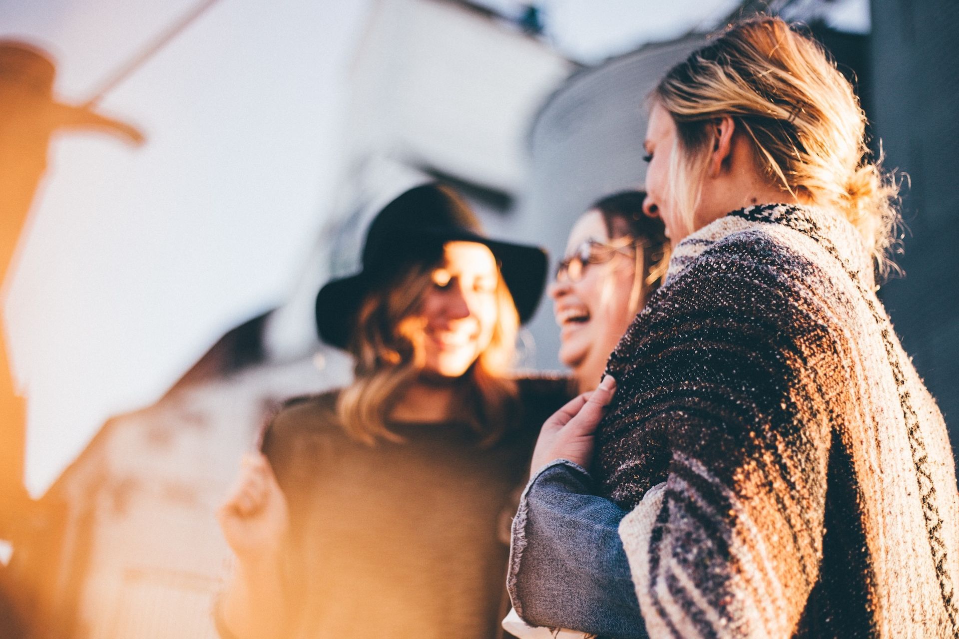 Three women laughing and embracing outdoors; one in a black hat, warm light.