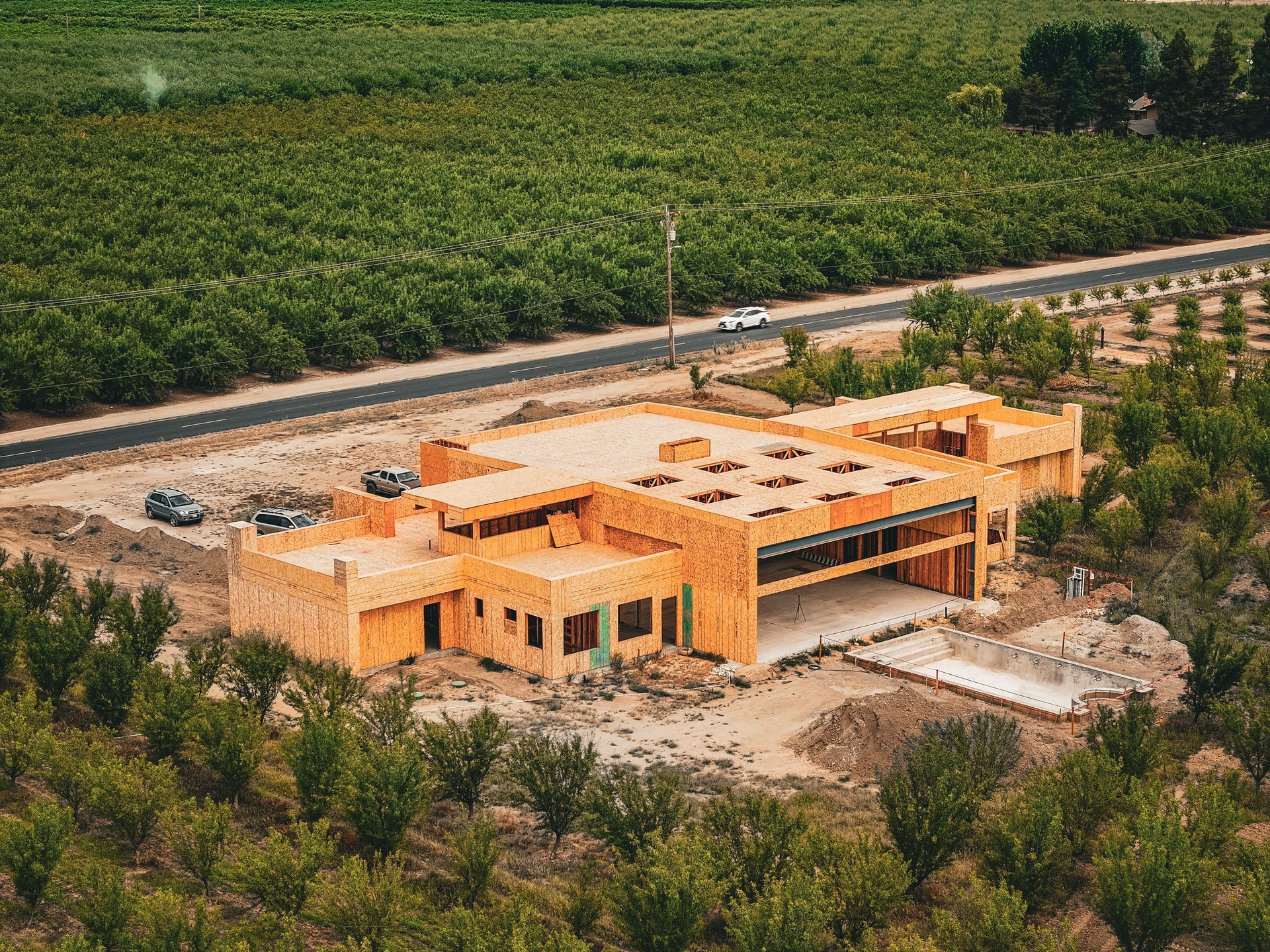 Aerial view of a wooden house under construction beside a road, surrounded by green fields and trees