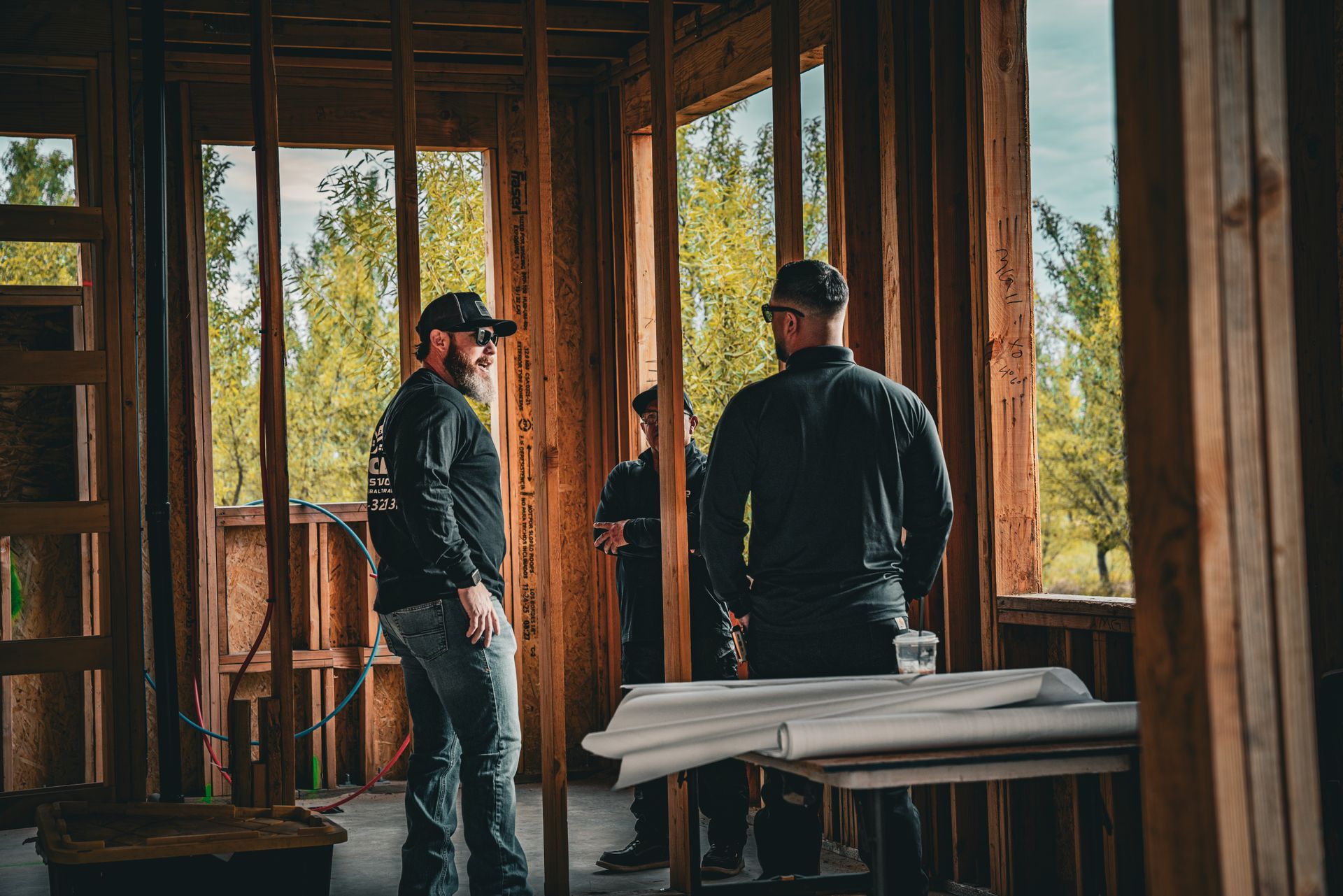 Two workers in dark jackets stand inside an unfinished wooden building with large open window frames.