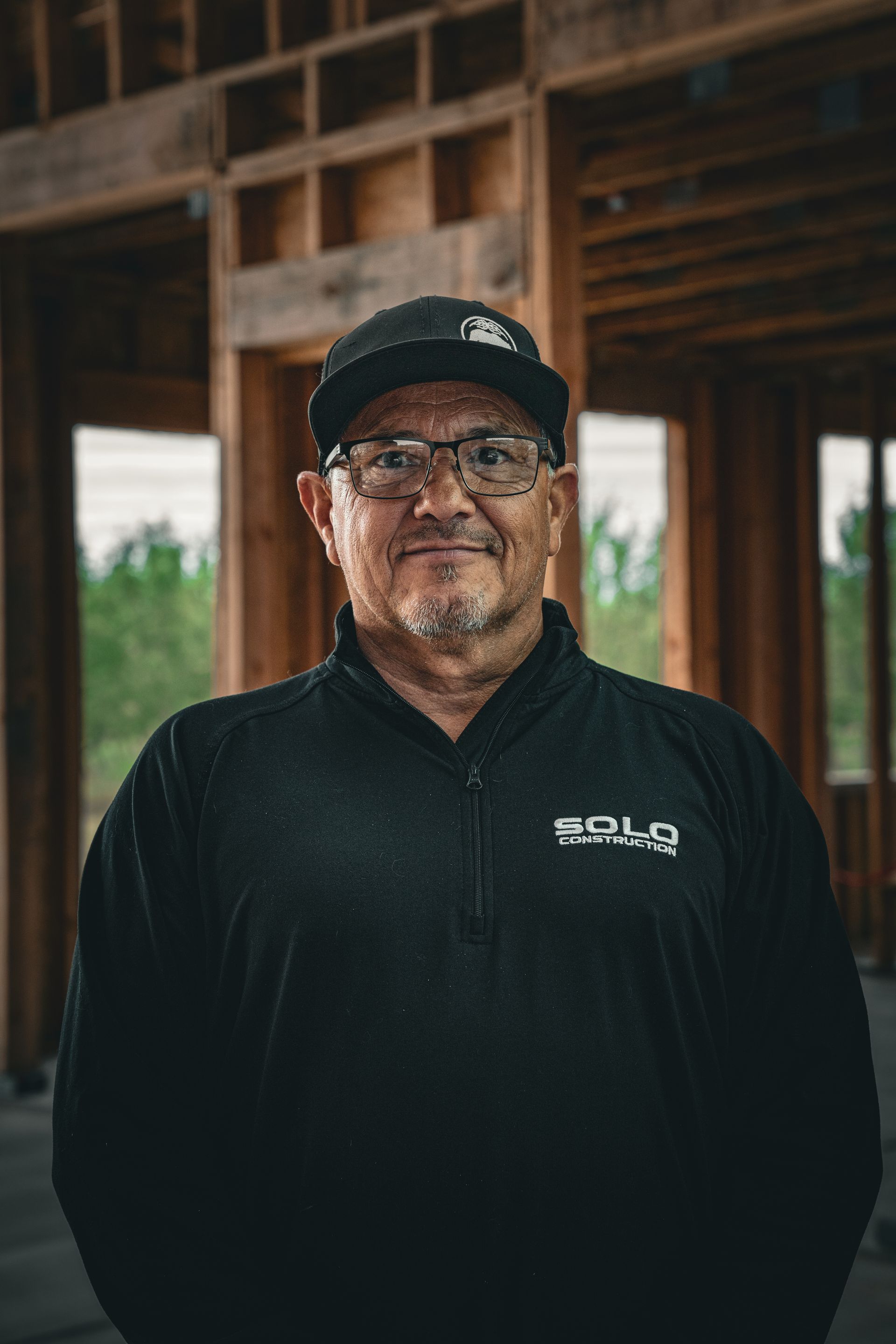 Man in black cap and shirt stands in a wooden structure, facing the camera with a neutral expression.