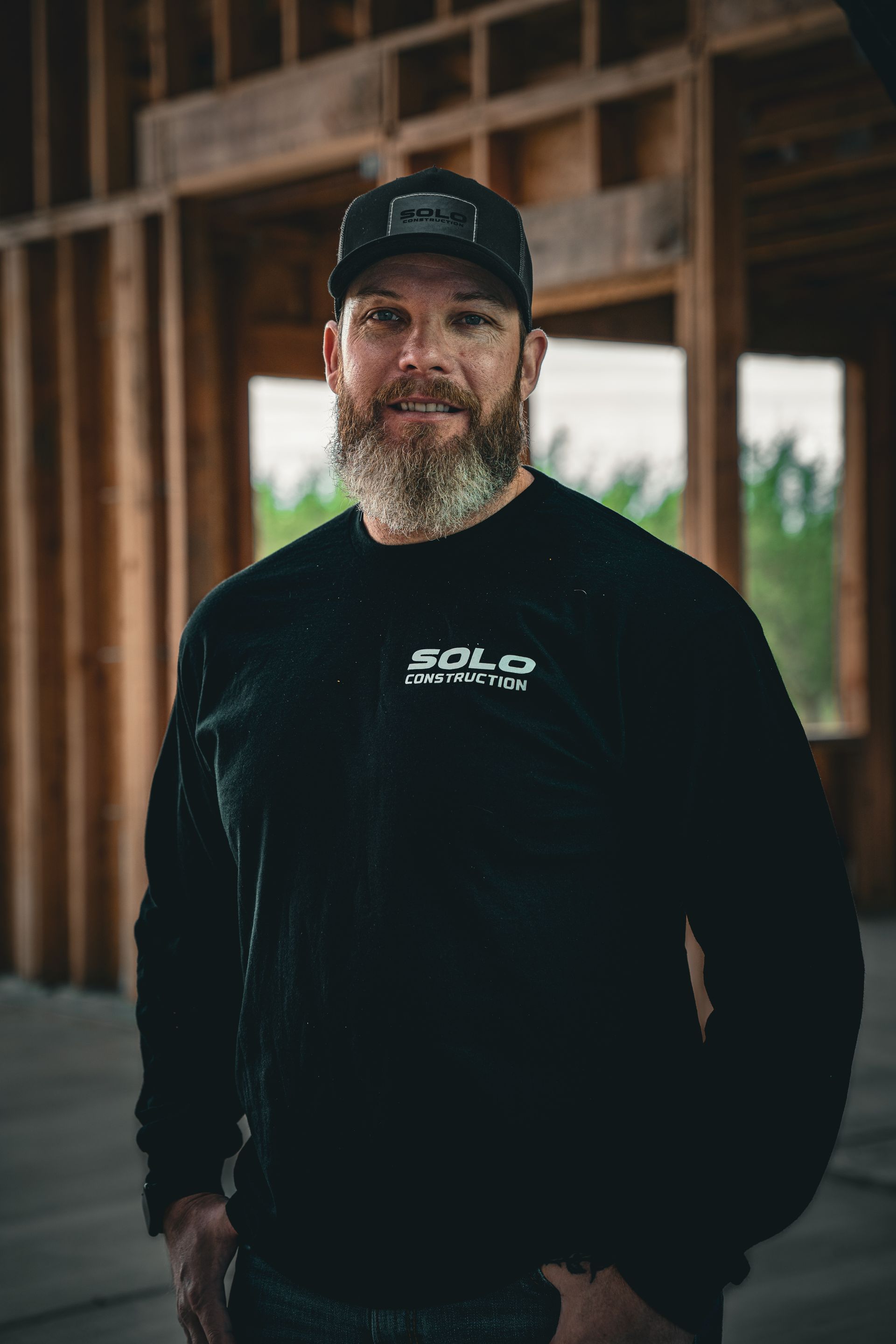 Man standing in a wooden structure, wearing a black cap and black shirt, with a beard and slight smile.