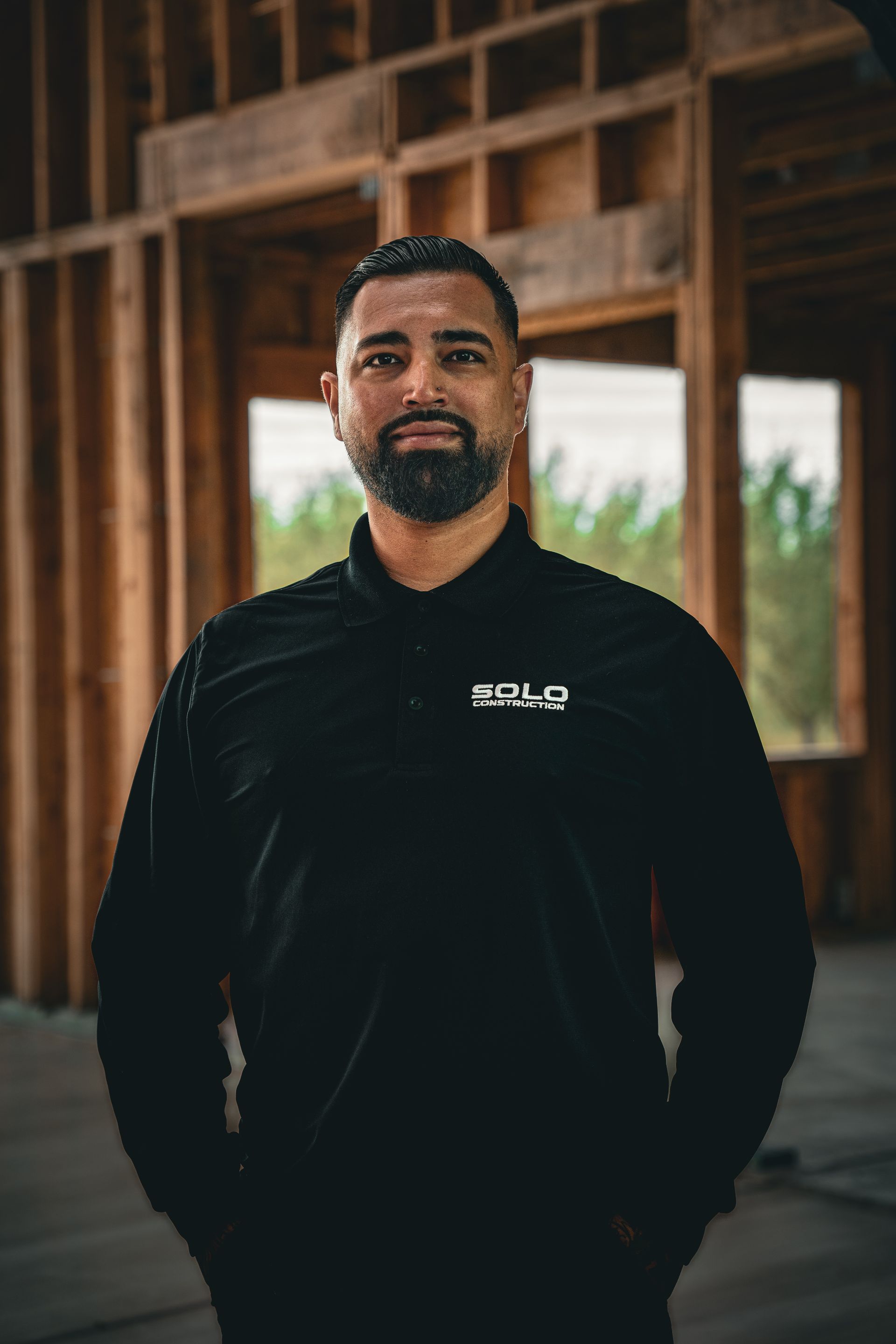 Man in black shirt standing in a wooden building, facing the camera with hands in pockets.