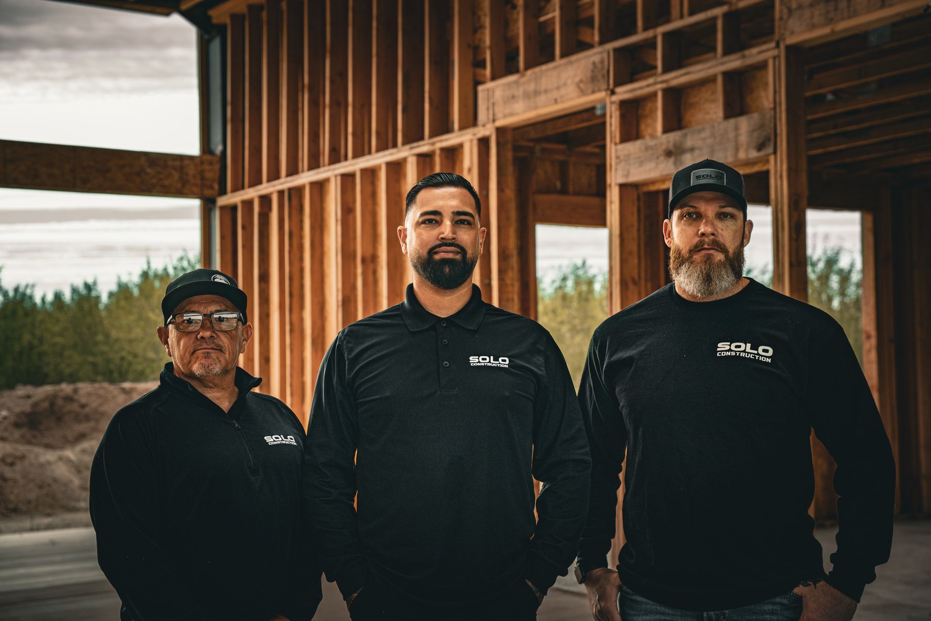 Three people in black jackets and caps standing in front of a wooden building under construction.