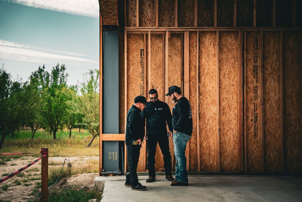 Three people stand talking beside a wooden building in an open outdoor area.