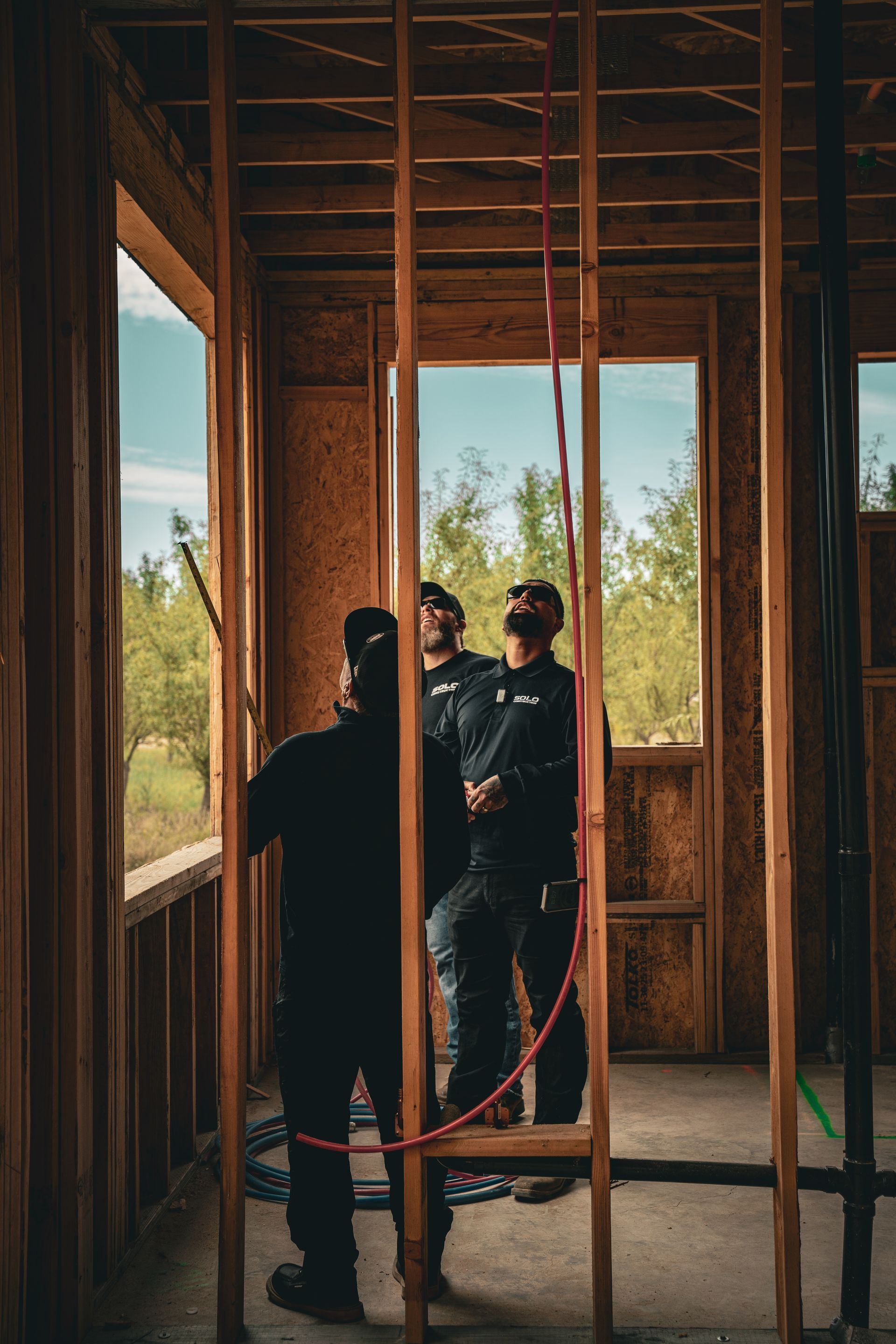 Three people in a wooden room under construction, looking out a window with trees outside.