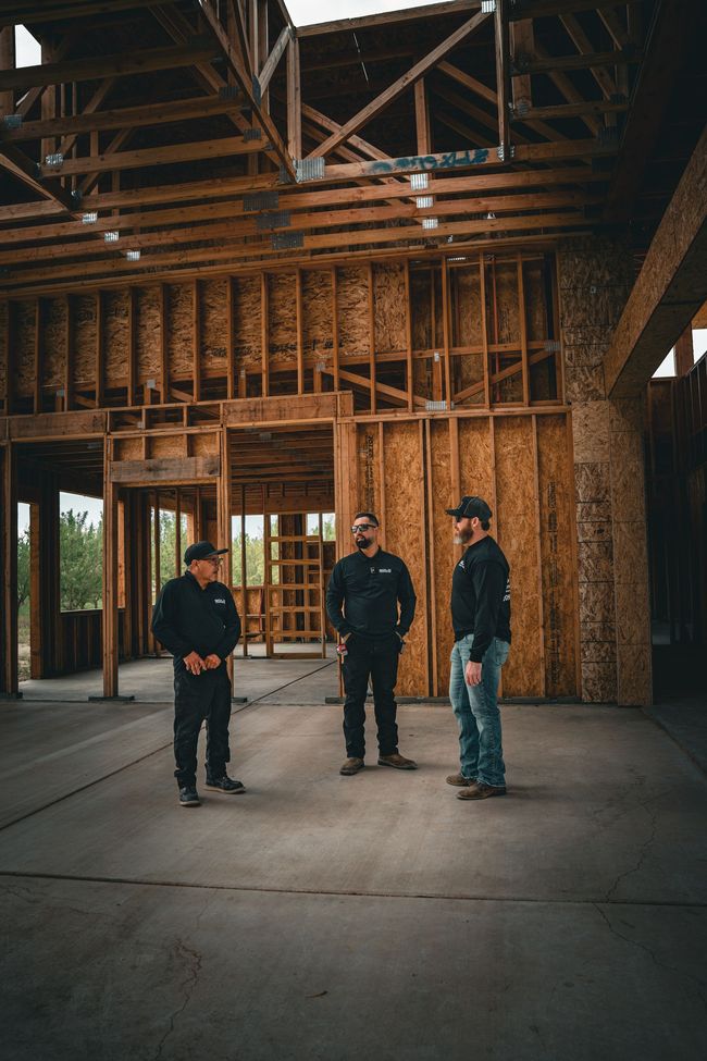 Three people stand inside an unfinished wooden building with exposed framing and an open doorway.