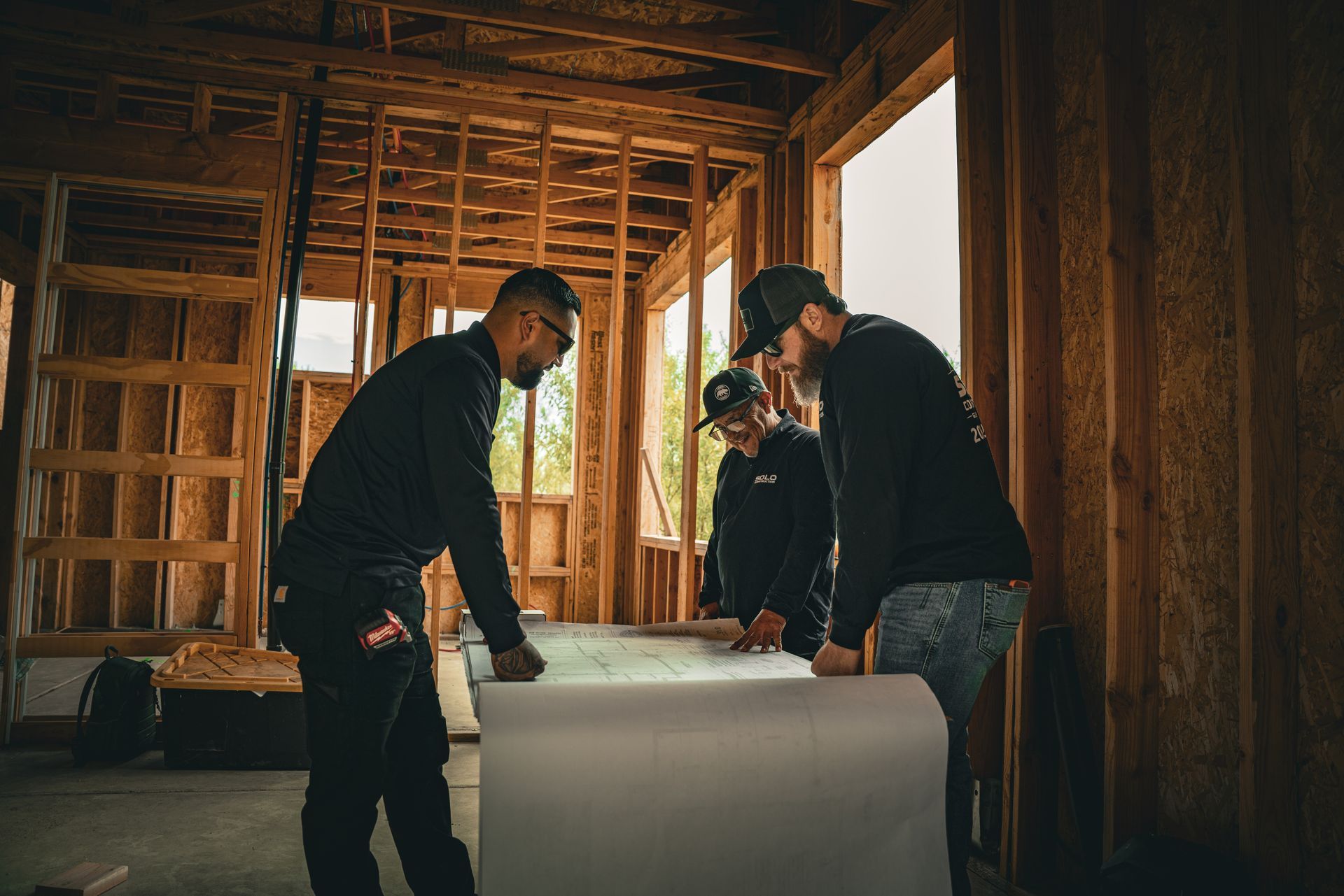 Three workers inspecting a large rolled-up sheet inside a wooden building under construction