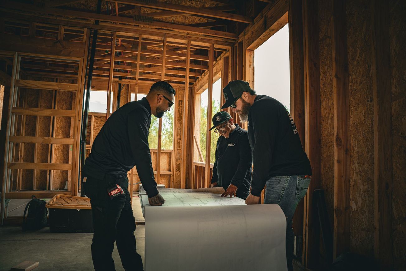 Two men in dark clothing stand in a wooden barn, one pointing upward while wearing a cap.