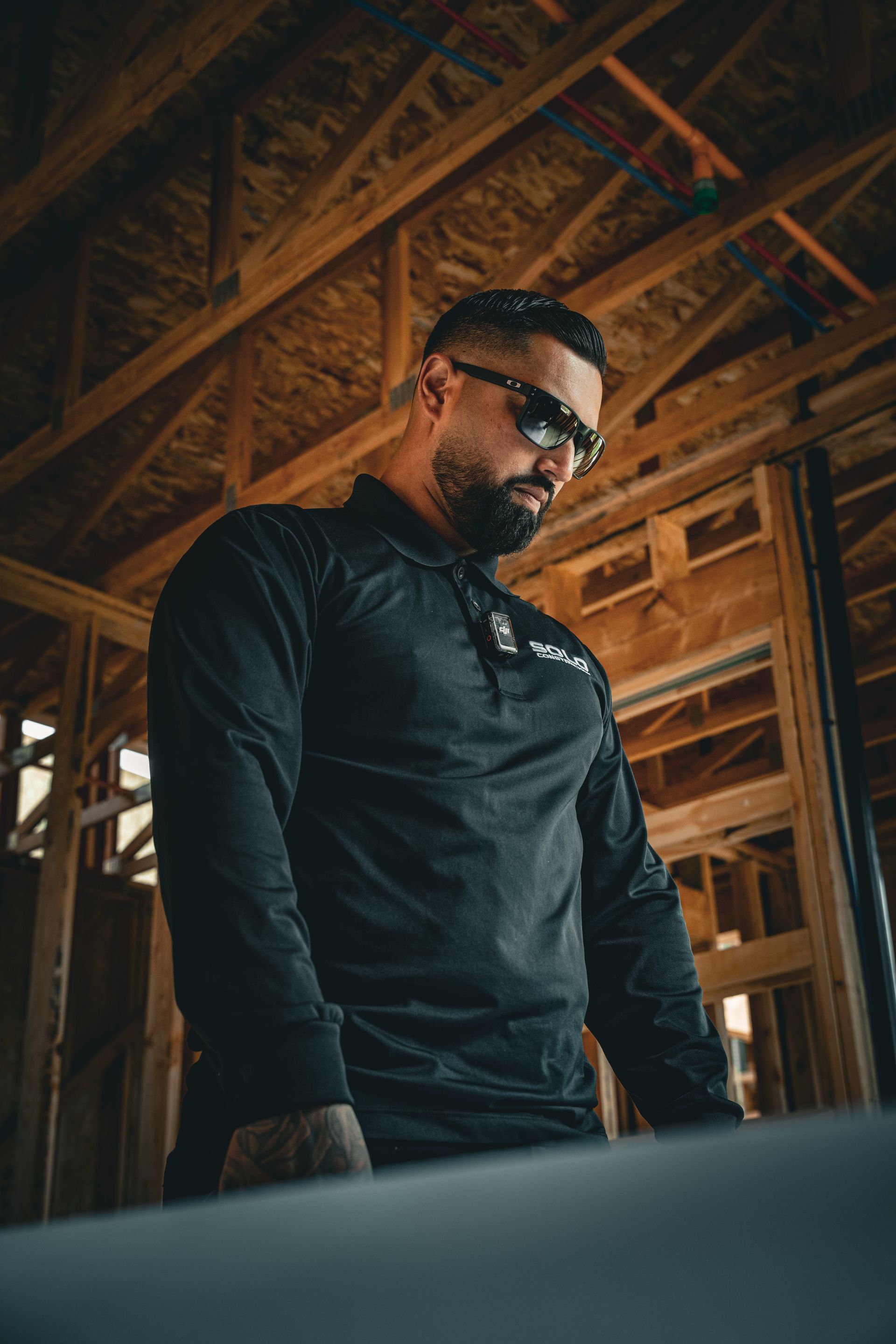 Man in black shirt and sunglasses standing in a wooden-framed construction site, looking down