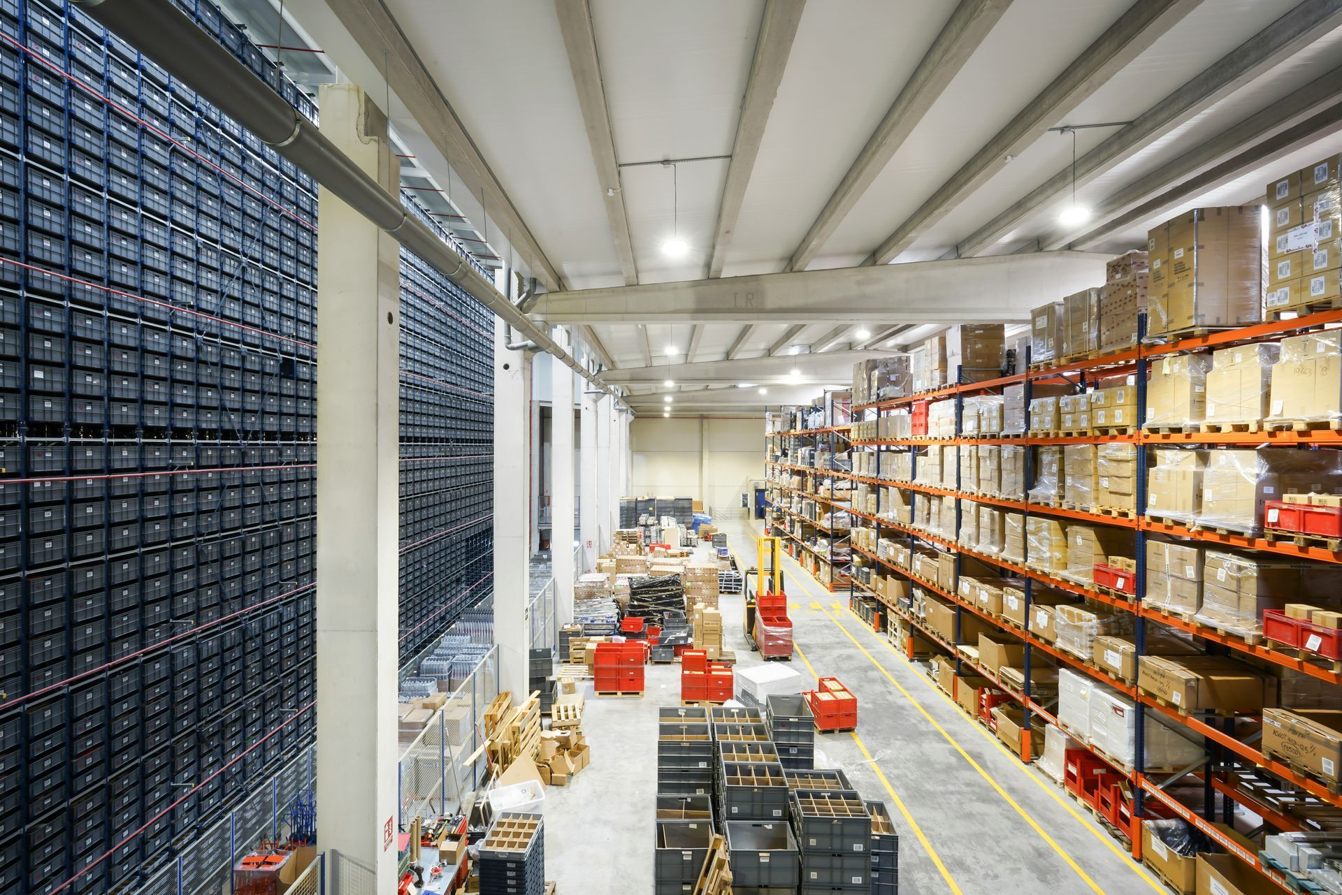 Warehouse aisle with stacked shelves, crates, and pallets under bright overhead lights