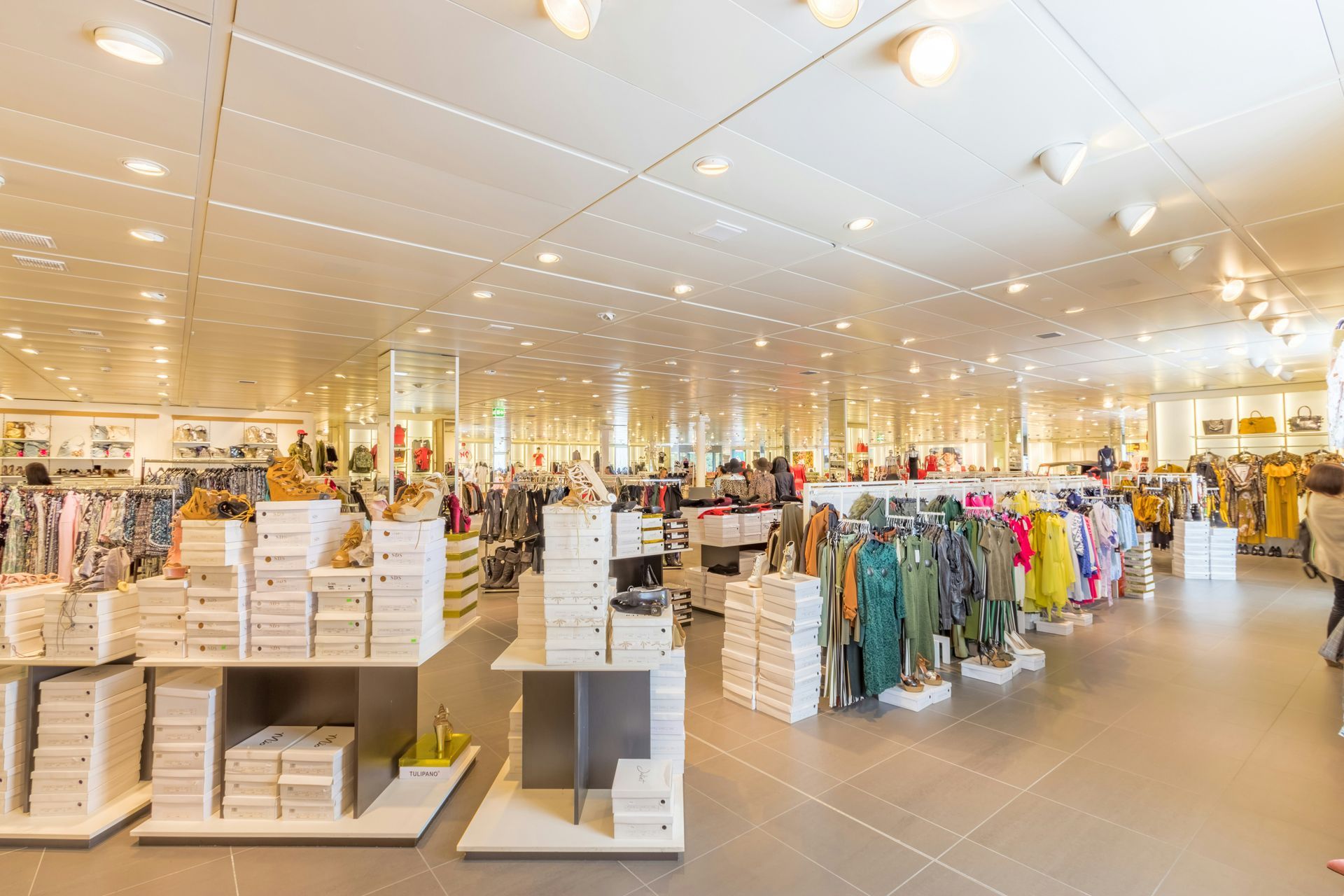 Bright retail store interior with shelves, clothing racks, and shoppers under warm ceiling lights