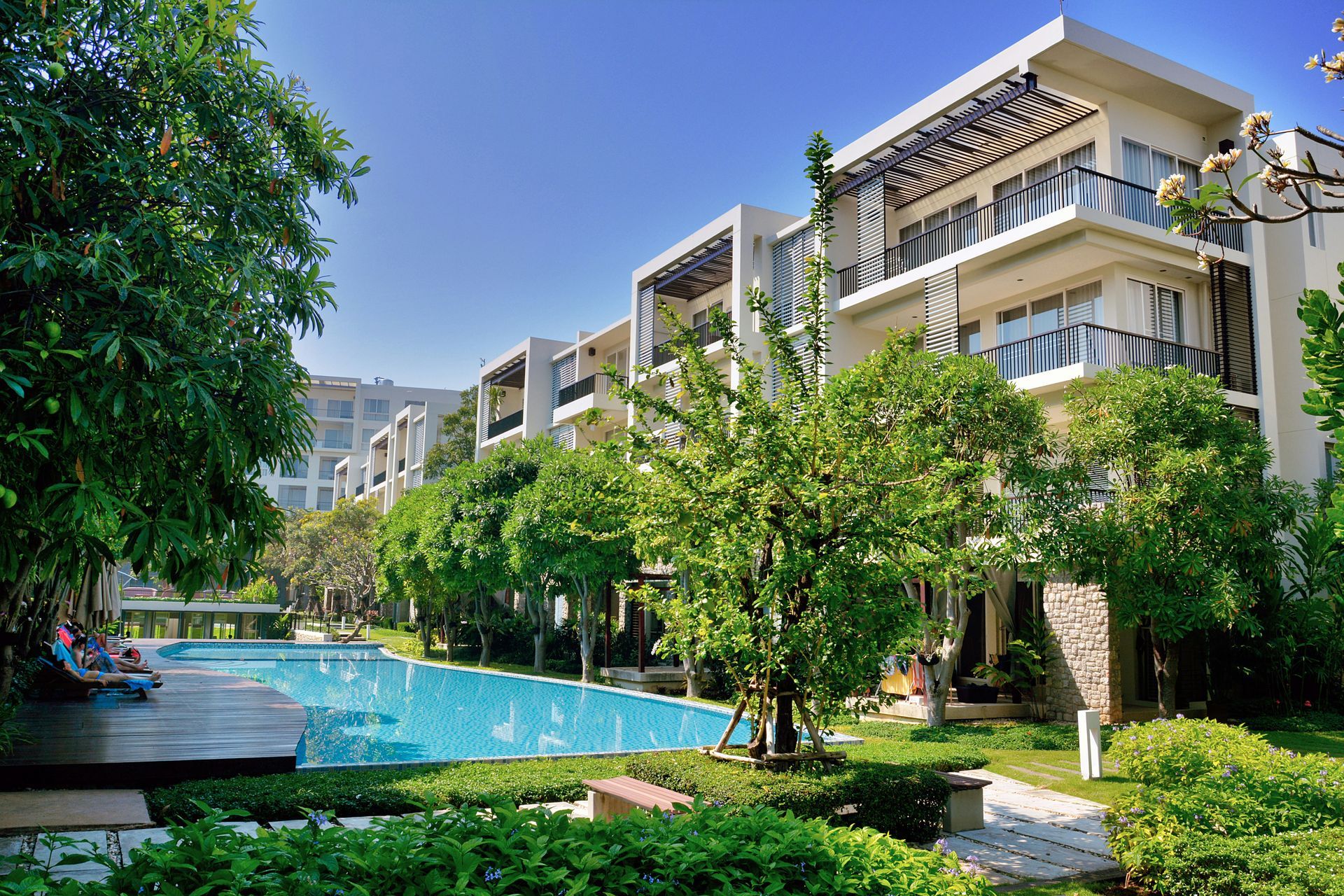 Modern apartment complex with balconies beside a blue swimming pool and lush green landscaping