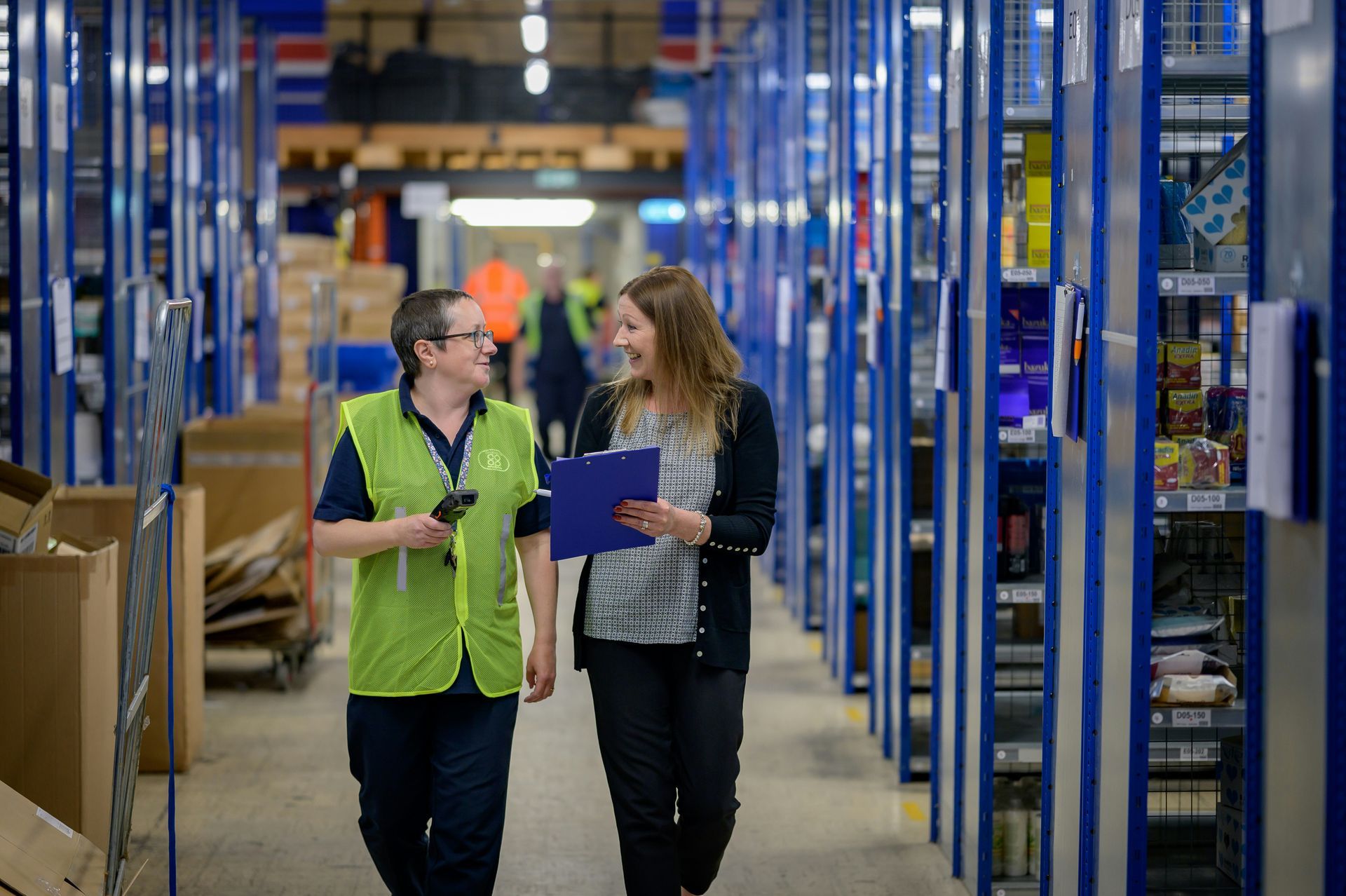 Two warehouse workers in high-visibility vests walk and talk between tall blue storage racks.