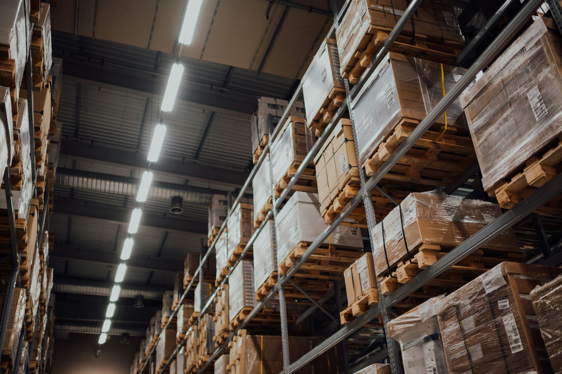 Warehouse aisle with tall pallet racks stacked with shrink-wrapped boxes and overhead lights