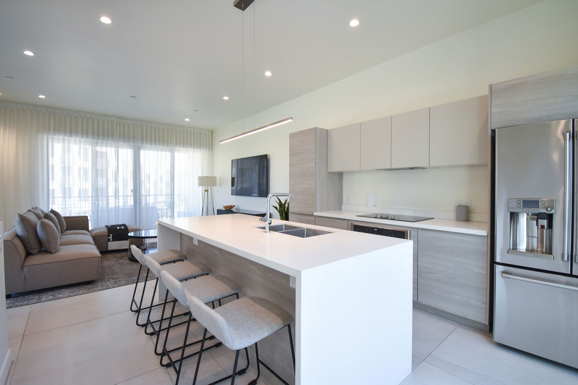 Modern open-plan kitchen and living room with white island, gray sofa, bar stools, and stainless steel fridge.