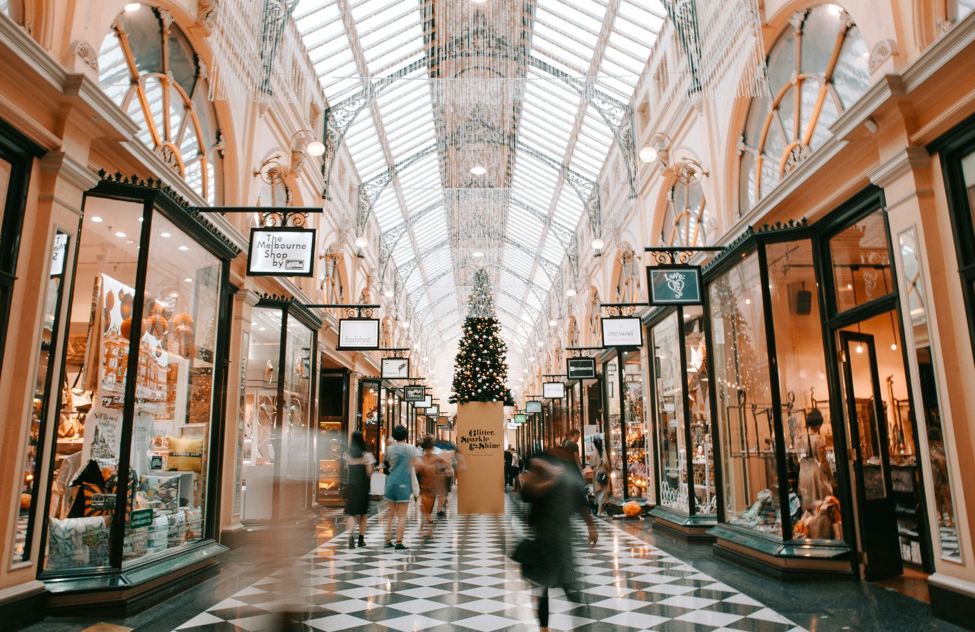 Elegant indoor shopping arcade with glass storefronts, checkered floor, and a decorated Christmas tree overhead