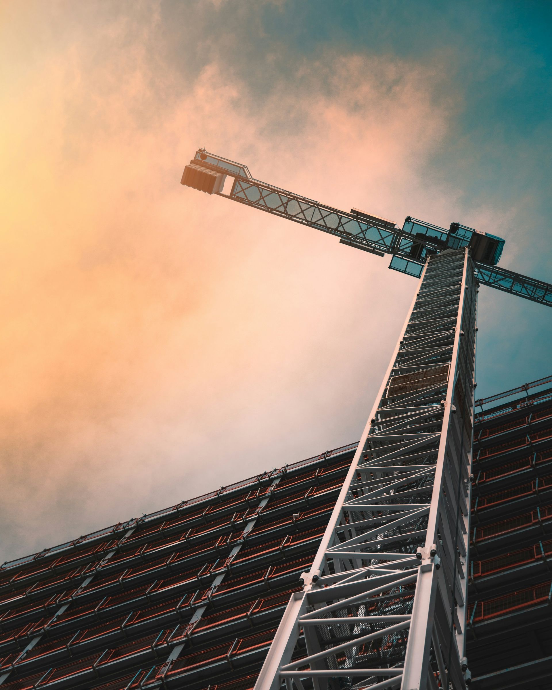 Low-angle view of a crane against a warm orange sky beside a dark building facade
