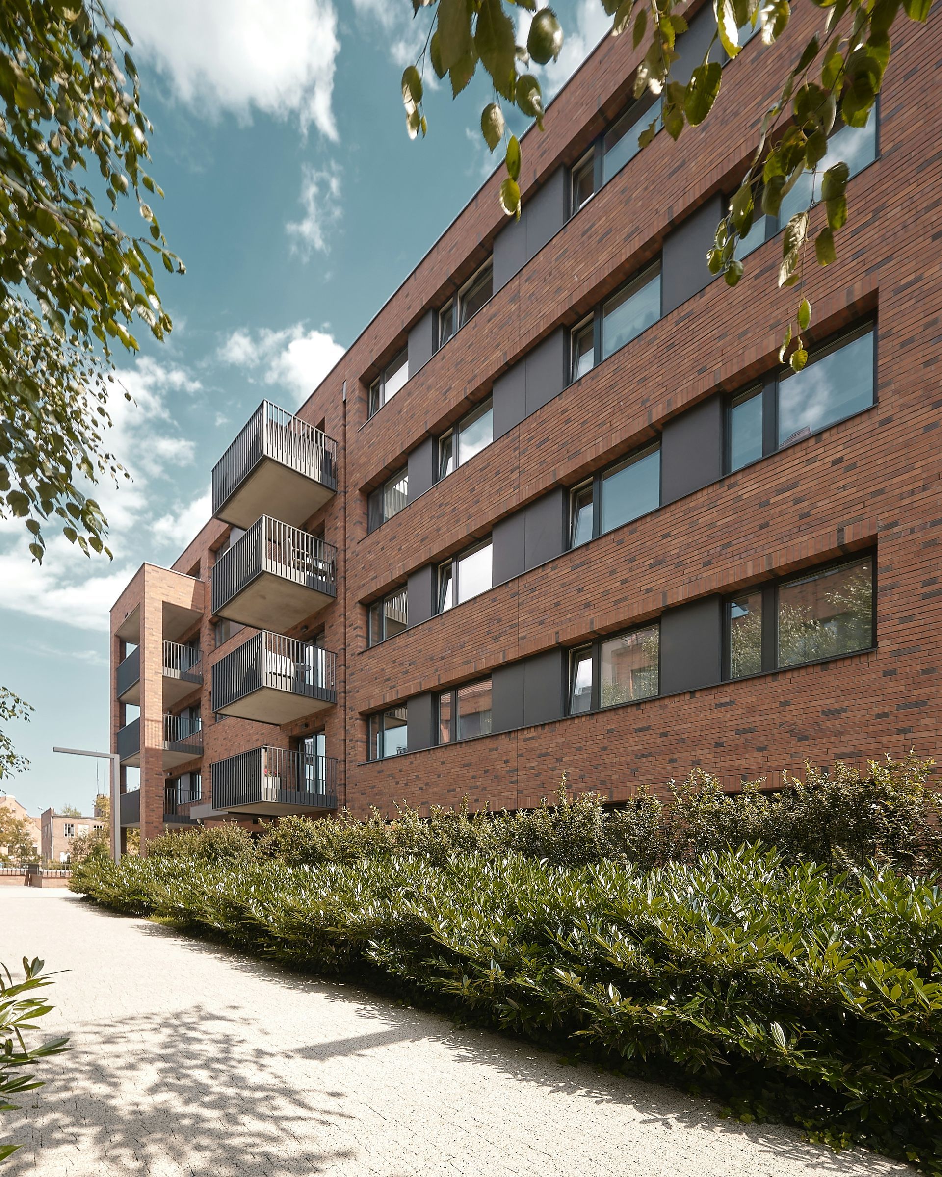 Modern brick apartment building beside a sidewalk and hedges on a sunny day