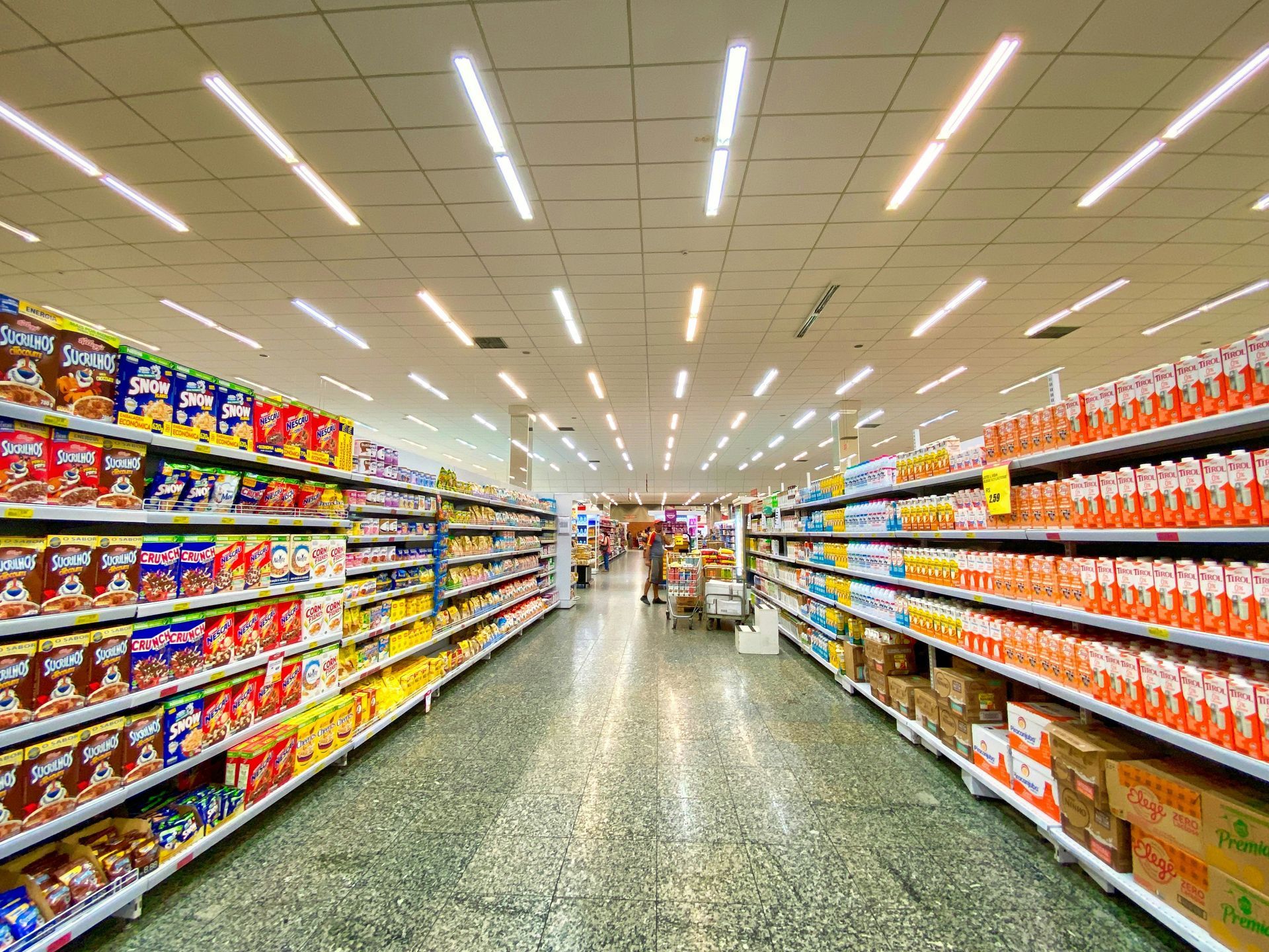 A brightly lit supermarket aisle with stocked shelves of colorful products on both sides.
