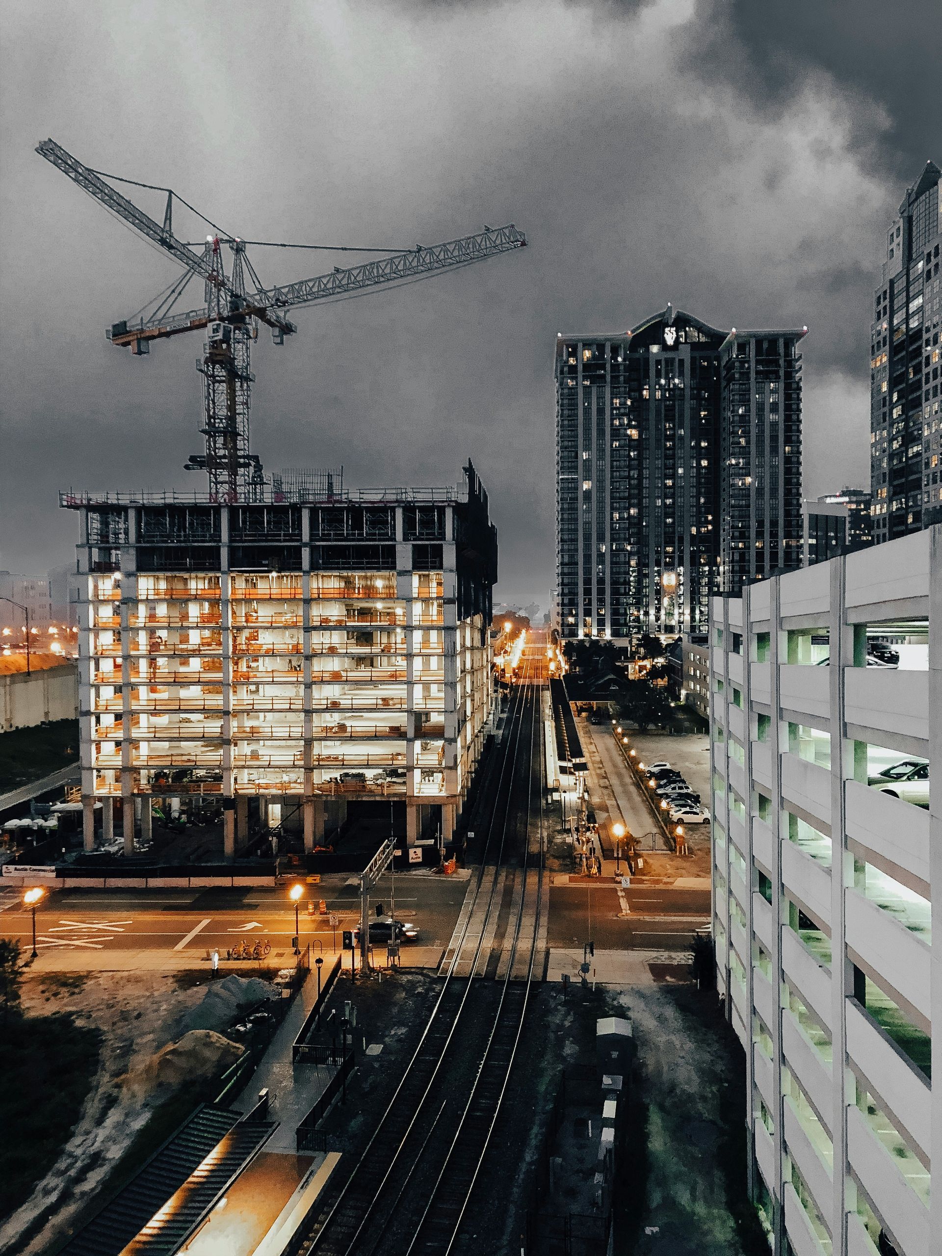 Night city construction site with crane, lit building, and cloudy sky beside a high-rise.