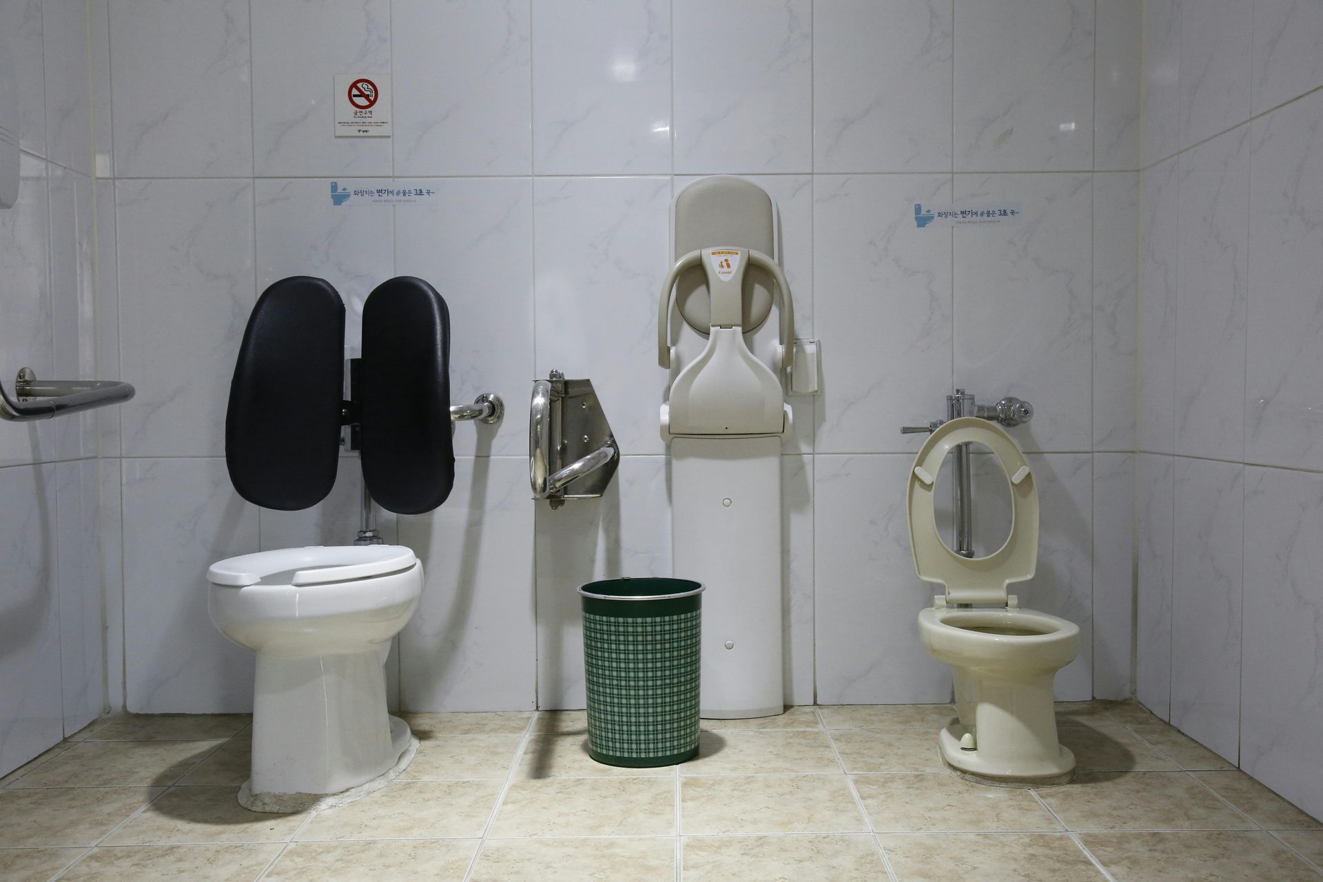 Public restroom with three toilets, one black-lidded and two cream-colored, against white tiled walls.
