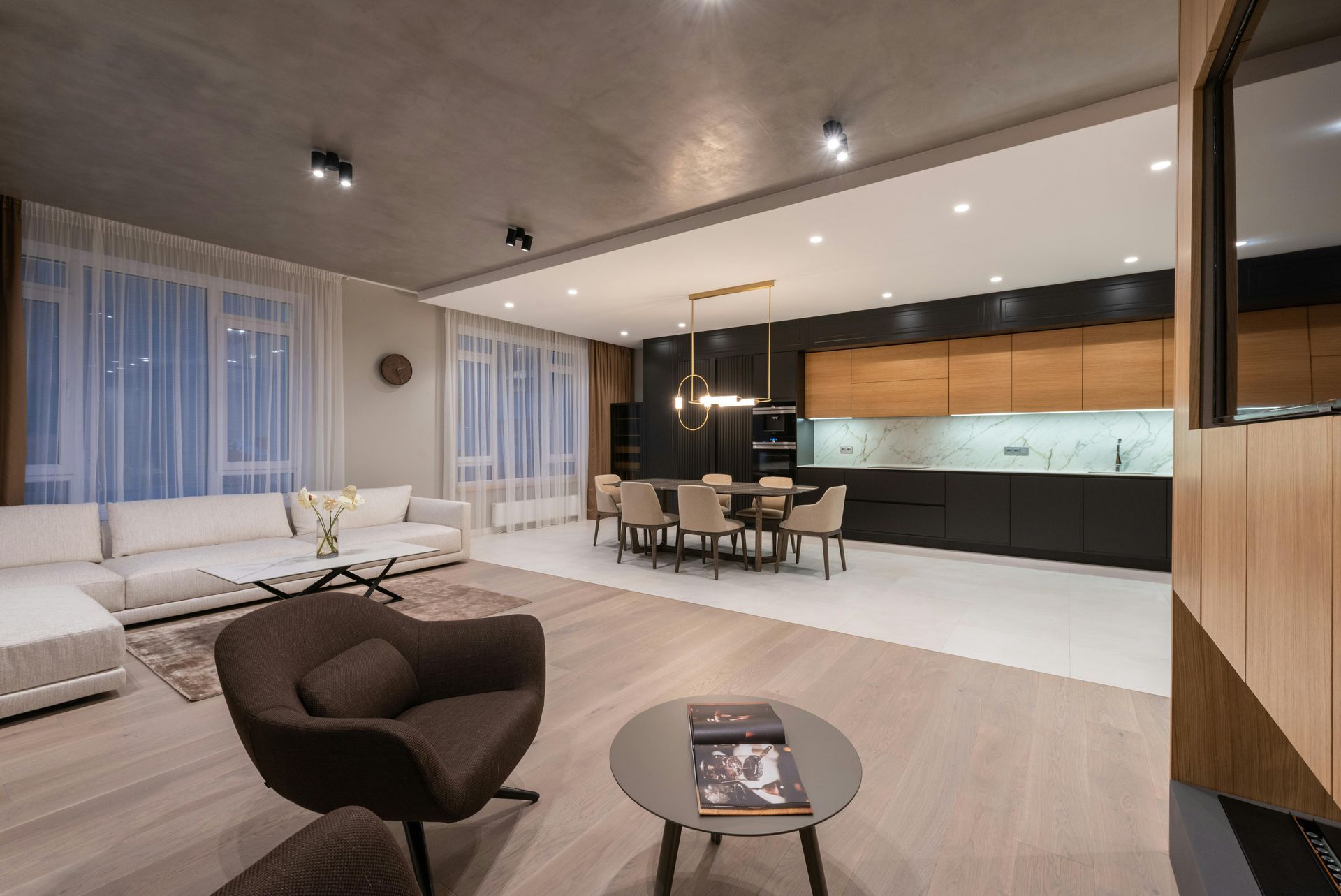 Modern open-plan living room and kitchen with neutral decor, white sofas, dark chairs, and warm wood accents