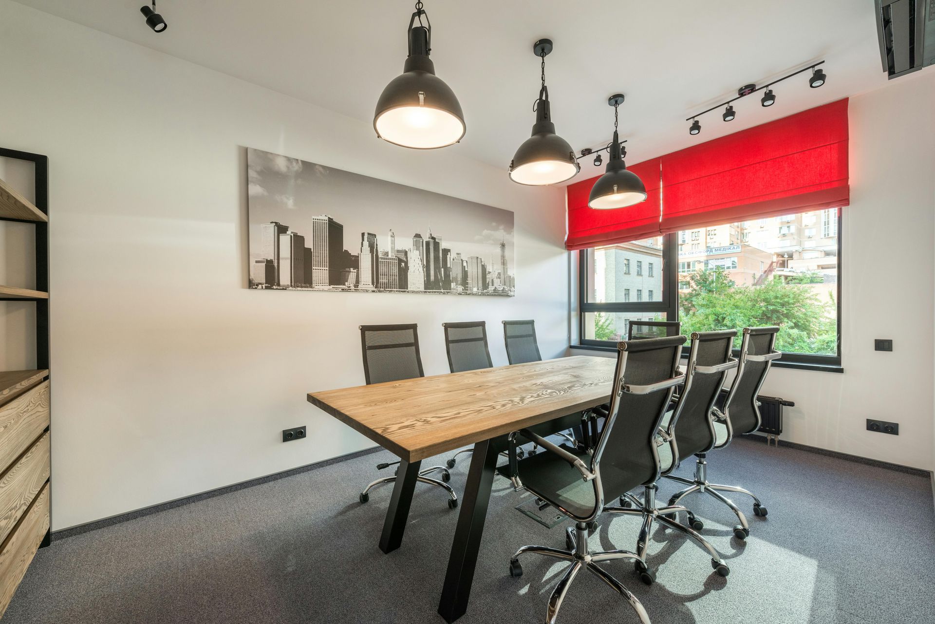 Modern conference room with wooden table, black chairs, pendant lights, city wall art, and a red window blind