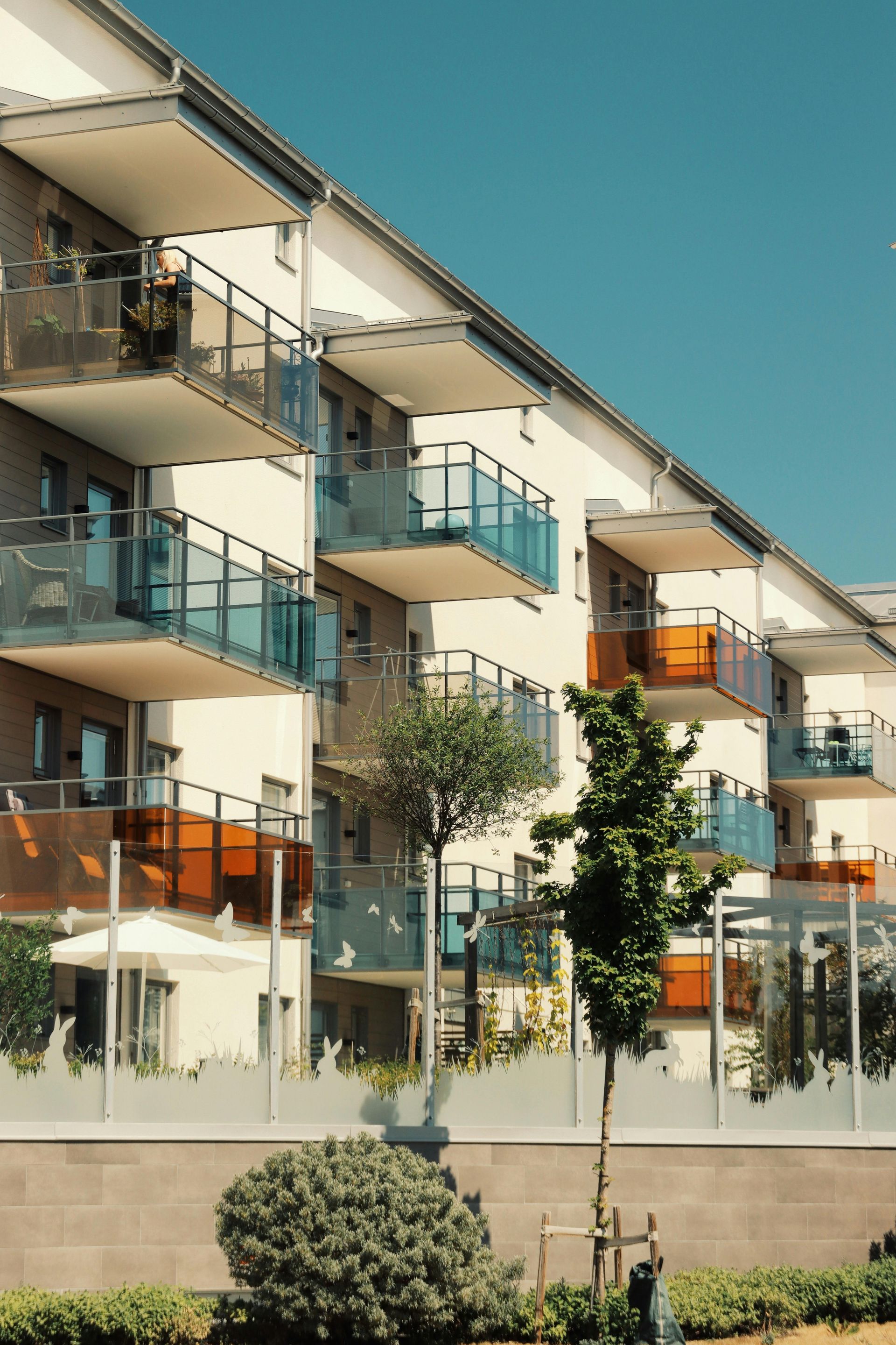 Modern apartment building with balconies, glass railings, and orange awnings behind a fence and shrubs