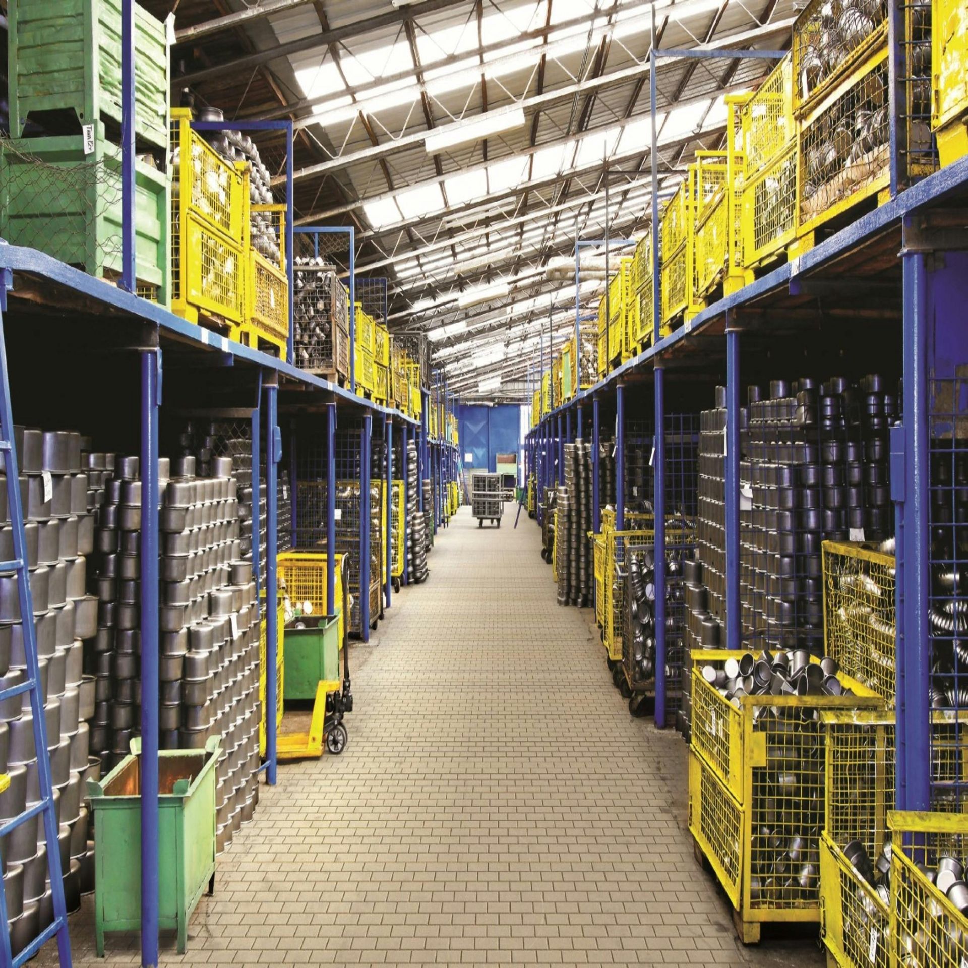 A warehouse aisle with stacked blue racks, yellow railings, and rows of metal barrels under a skylit roof