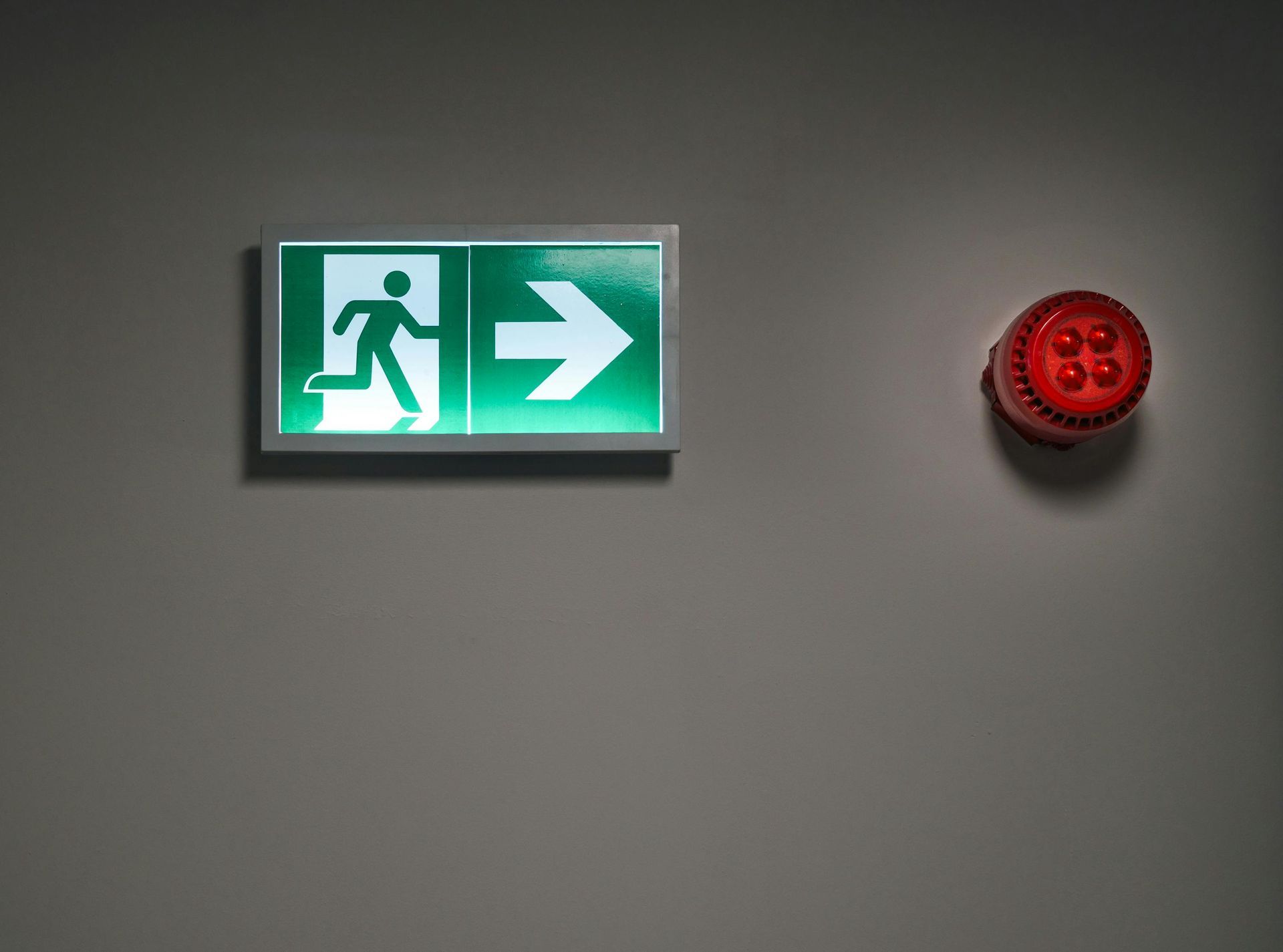 Green emergency exit sign with running figure and arrow beside a red alarm on a gray wall