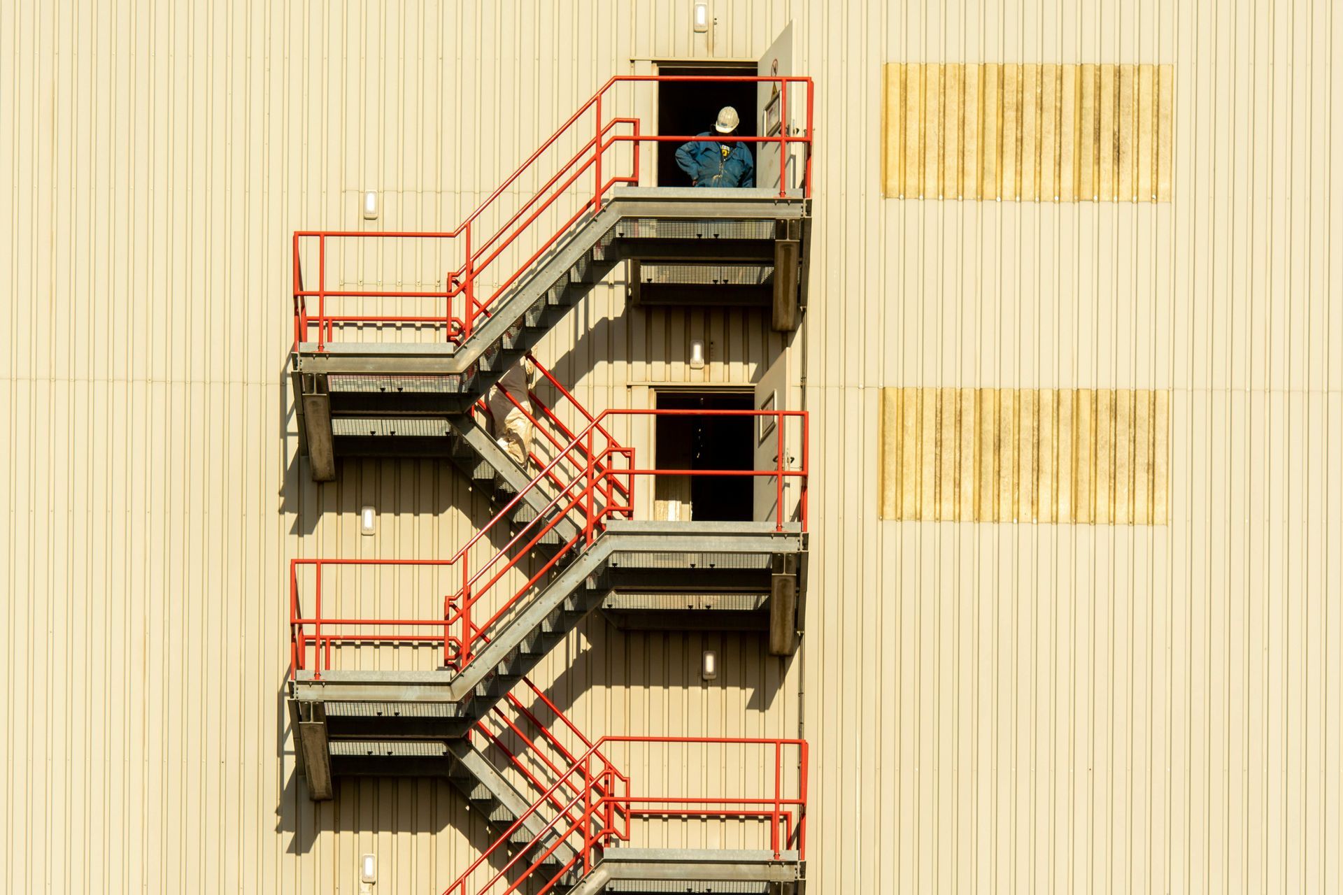 Red metal fire escape stairway on a beige building facade, with a person standing at the top landing.