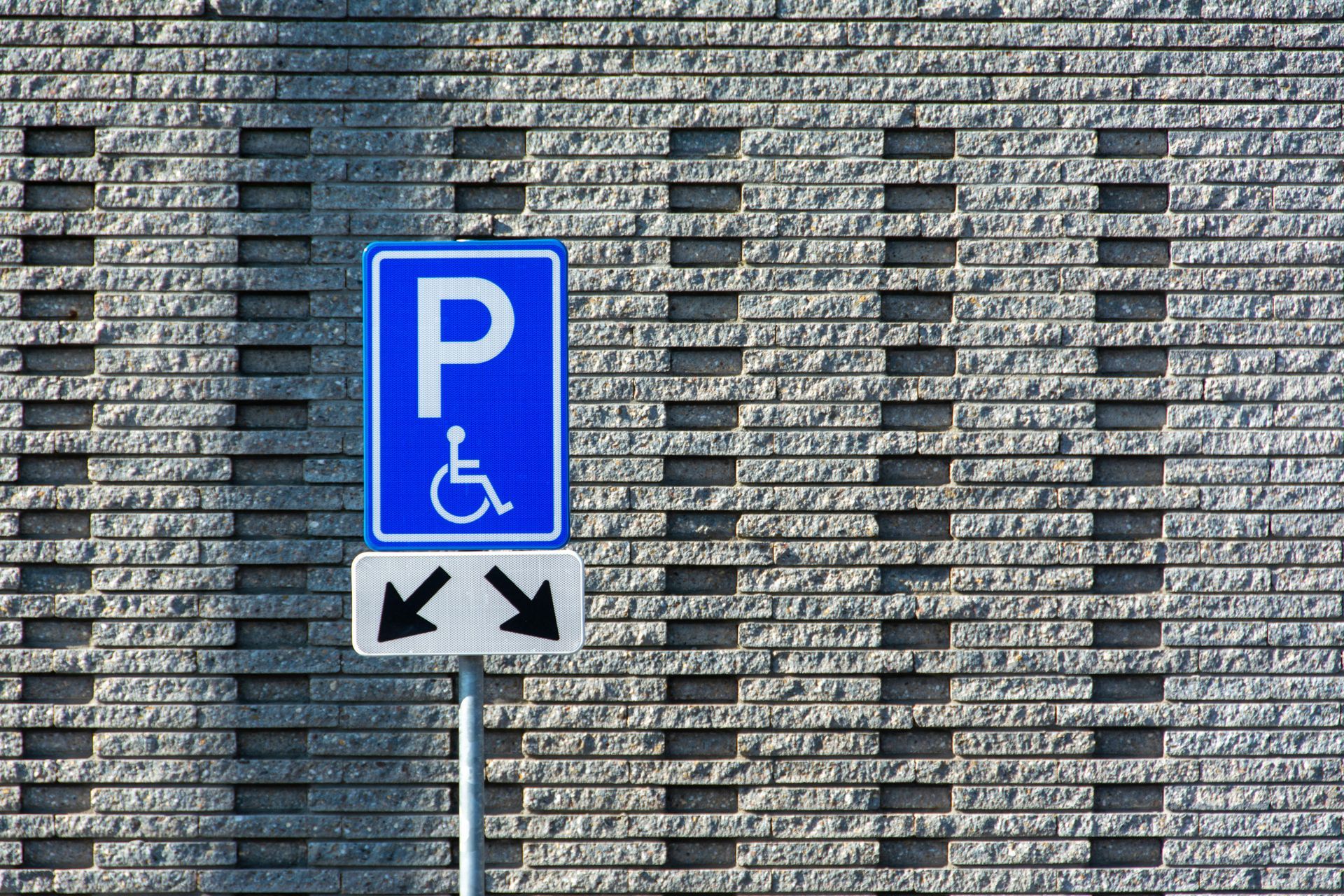Blue accessible parking sign with wheelchair symbol and directional arrows on a pole against a brick wall