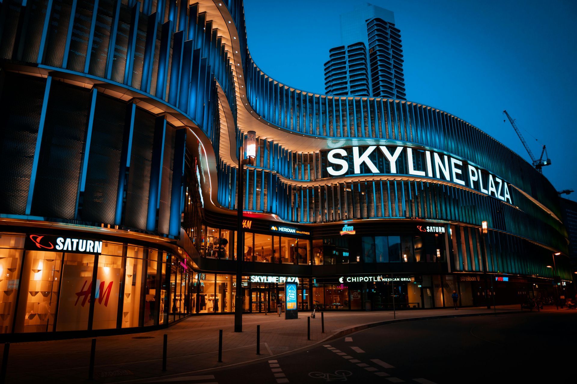 Night view of Skyline Plaza mall with glowing curved facade and blue-lit high-rise tower behind it