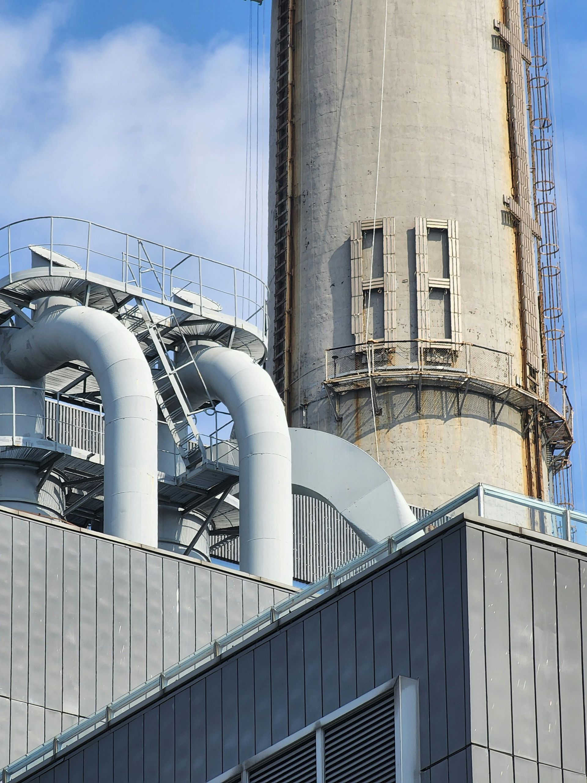 Industrial smokestack with large white pipes against a blue sky above a factory building