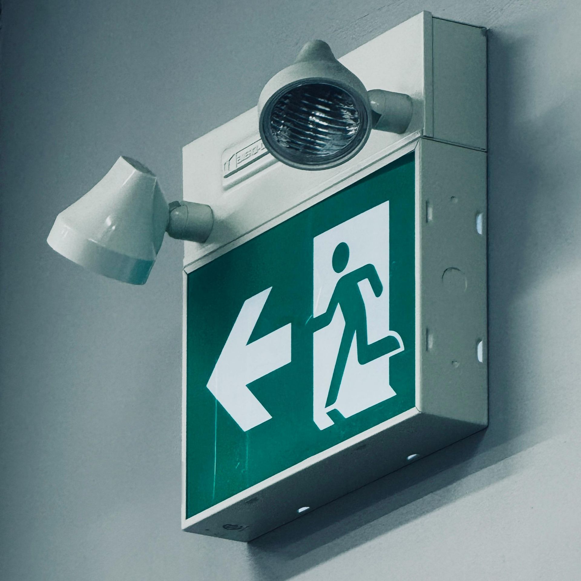 Emergency exit sign with left arrow and running figure, illuminated by a wall-mounted light
