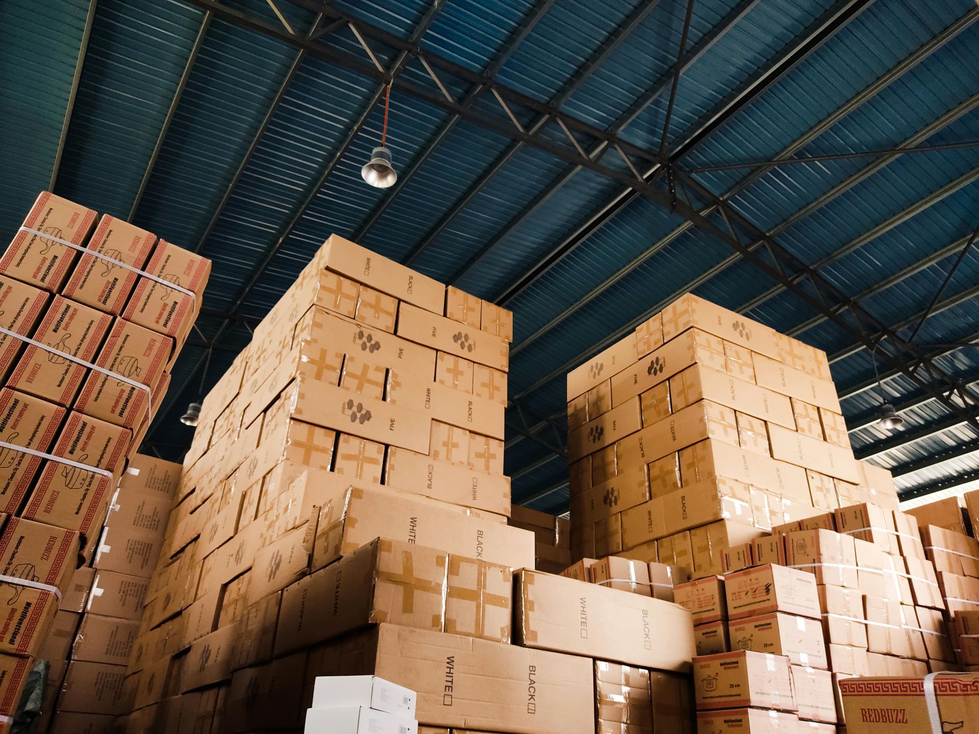 Stacks of cardboard boxes piled high in a warehouse with a blue metal ceiling.