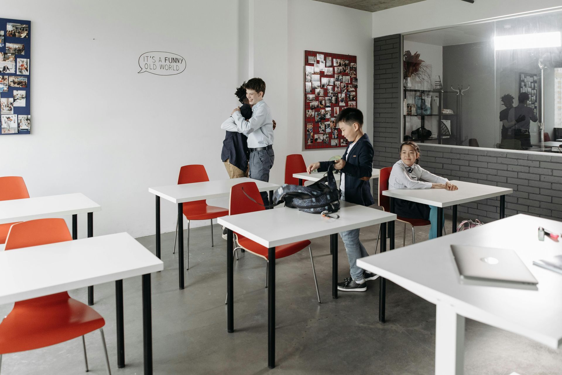 Classroom with white tables and red chairs; three people near the back wall and a counter area.