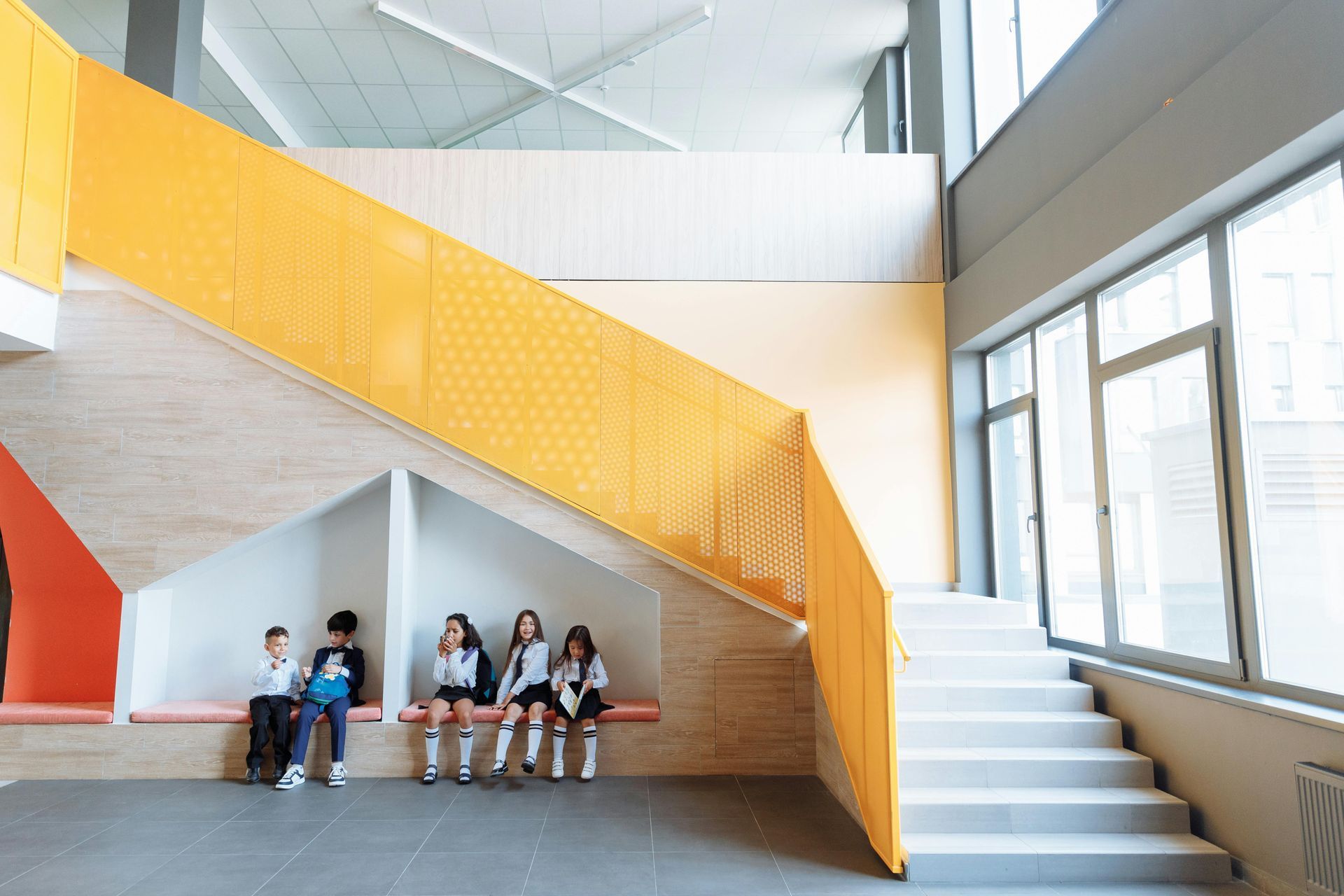 Students sitting under a yellow staircase in a bright modern school hallway