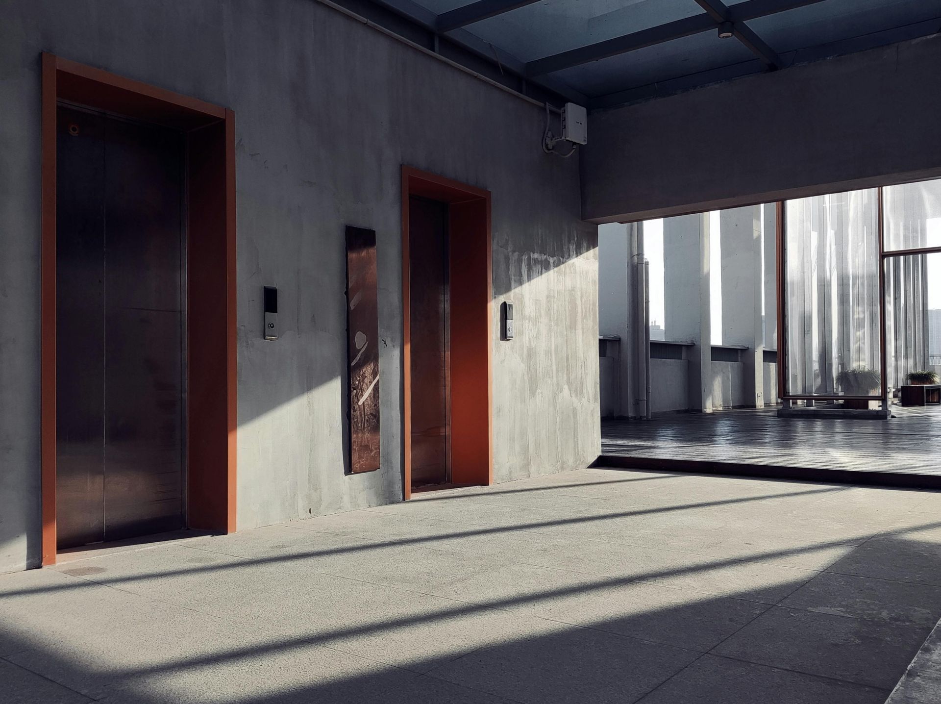 Modern building entrance with two elevators, concrete walls, and sunlit shadows on the ground.