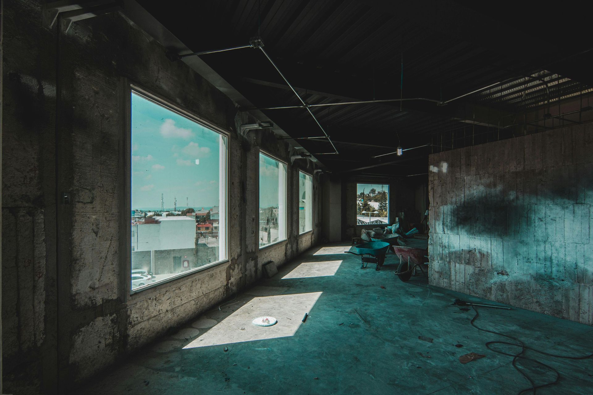Abandoned room with broken windows, peeling walls, and a blue-green floor flooded by daylight