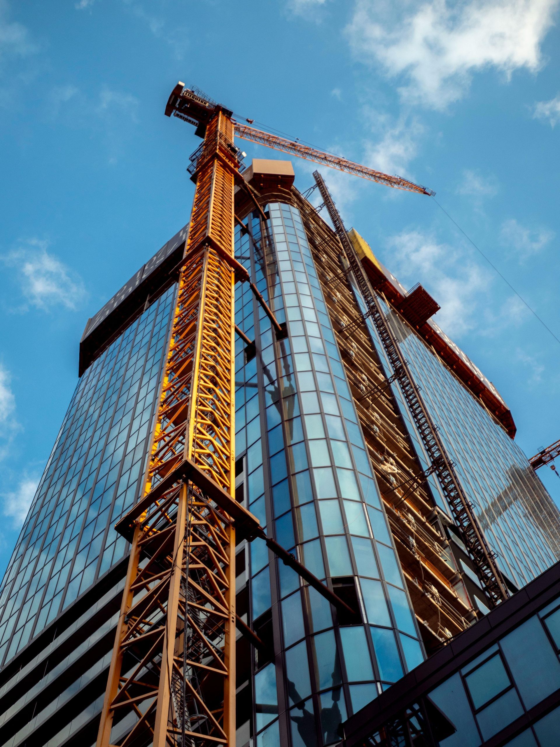 Tall glass office tower under construction with orange cranes against a blue sky