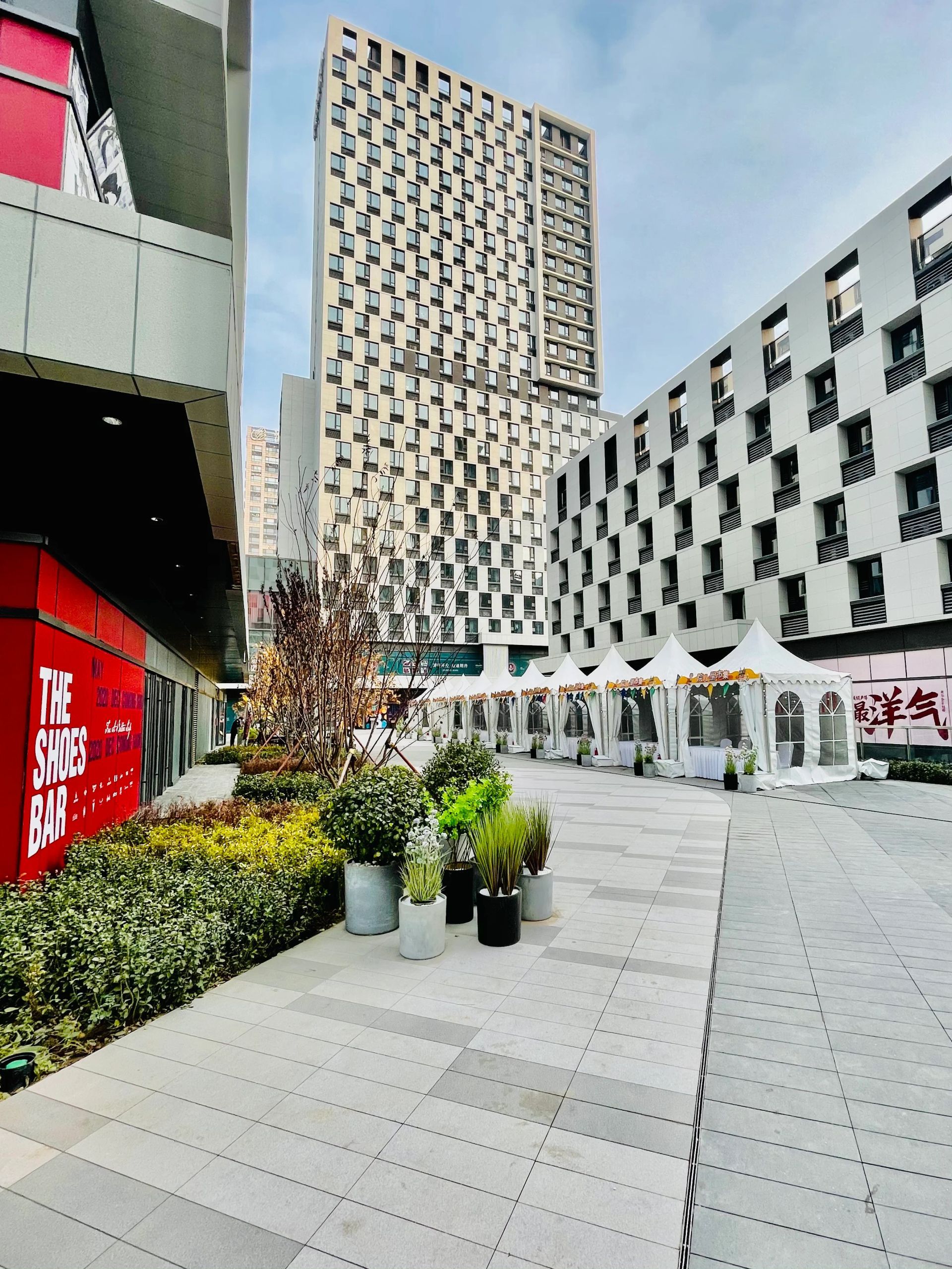 Modern plaza with geometric buildings, white tents, and potted plants under a bright sky
