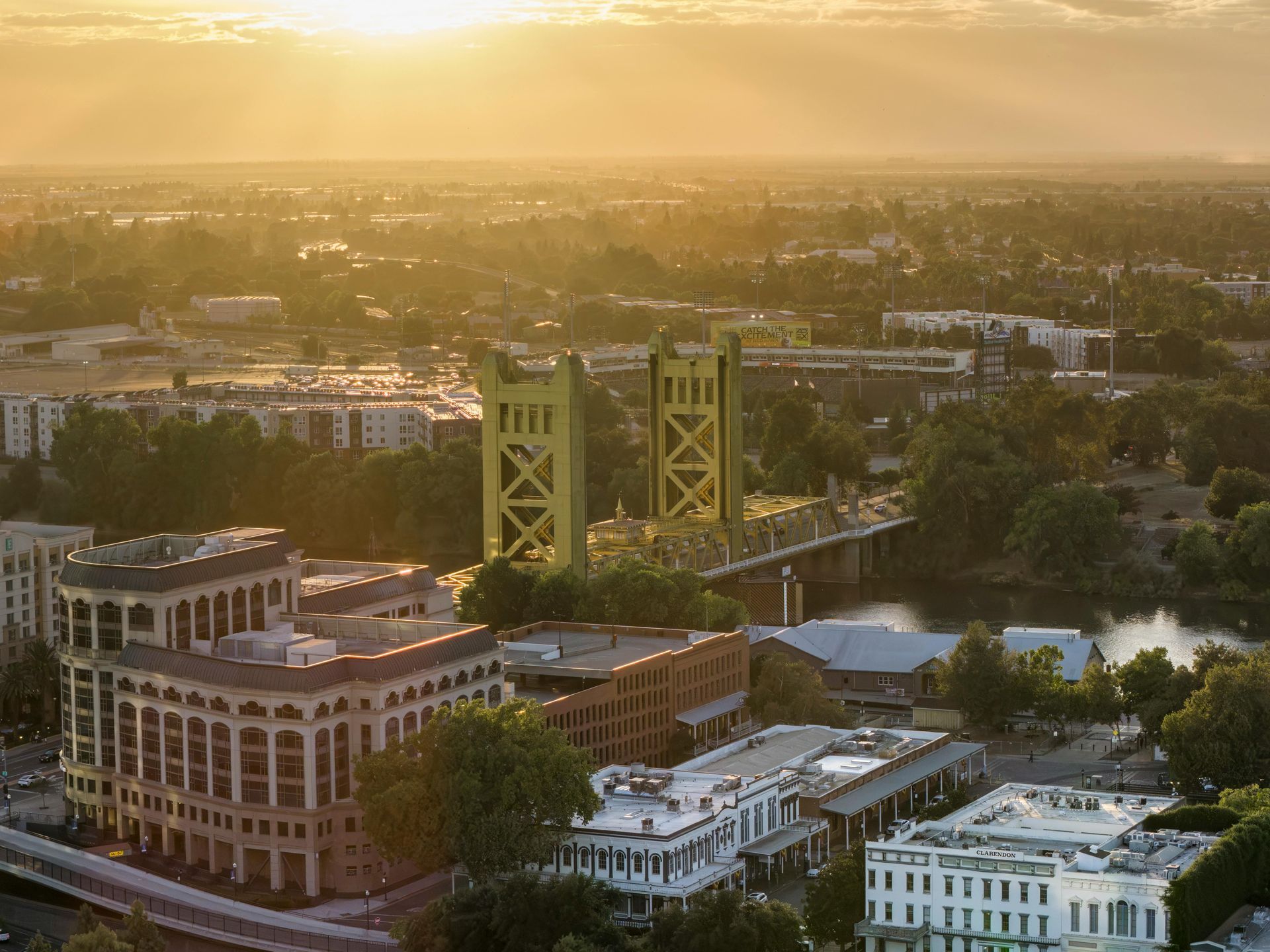Golden-hour cityscape with a green suspension bridge over a river and brick buildings in the foreground