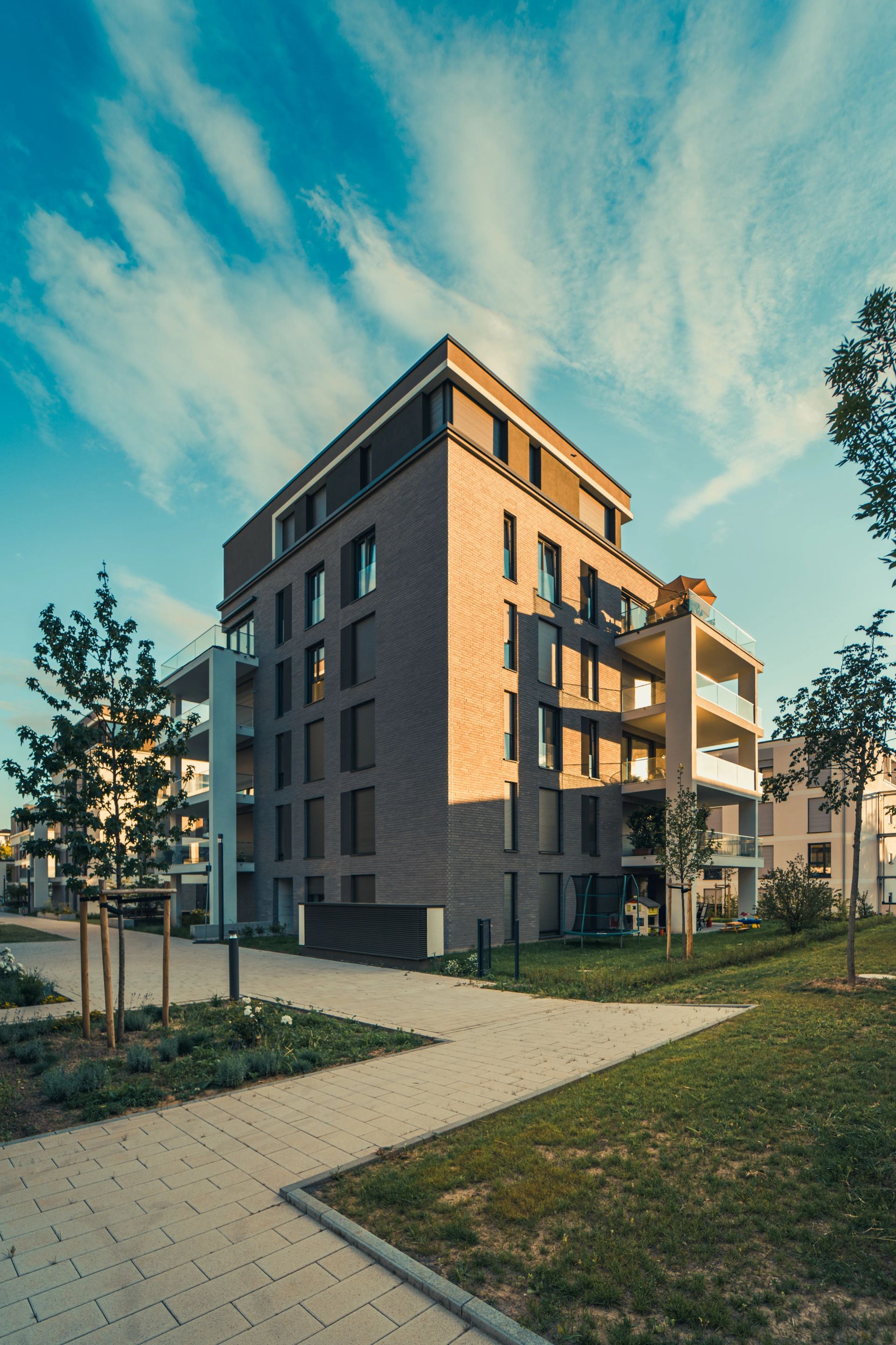 Modern brick apartment building at sunset, with balconies, trees, and a paved path in front.