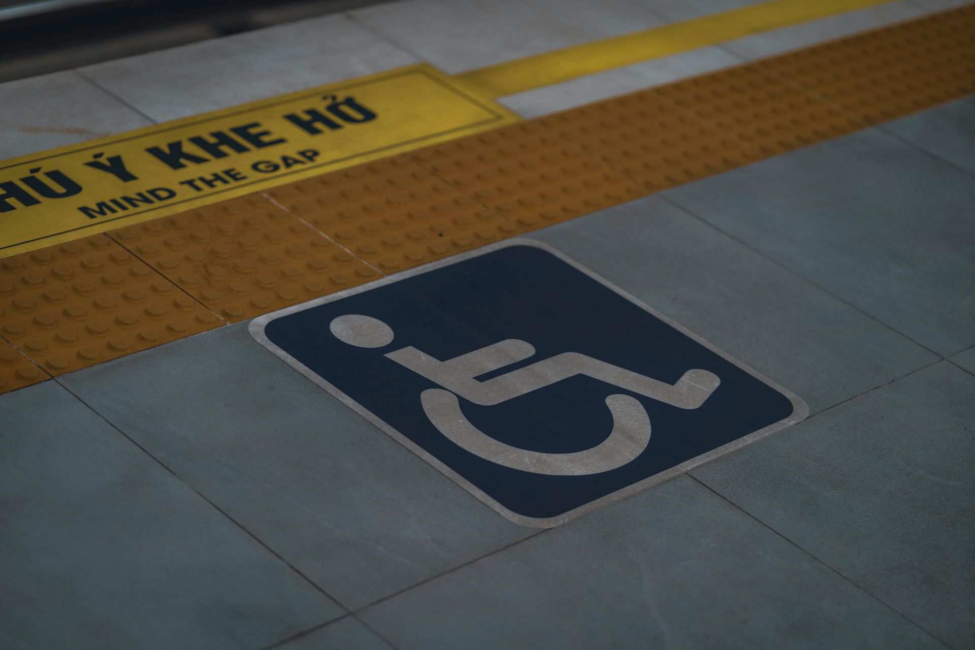 Blue wheelchair accessibility symbol painted on a tiled floor beside a yellow station sign.